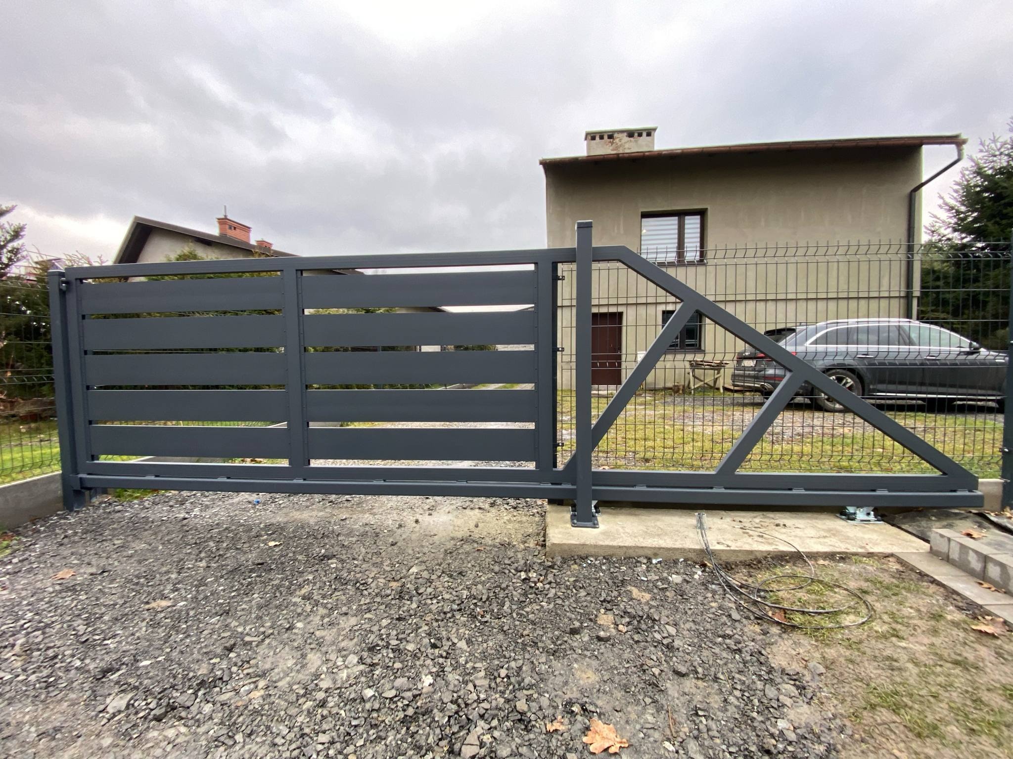 Nowoczesna, przesuwna brama wjazdowa z poziomymi panelami w kolorze grafitowym na tle domu i ogrodzenia panelowego. Widoczne detale konstrukcyjne bramy.