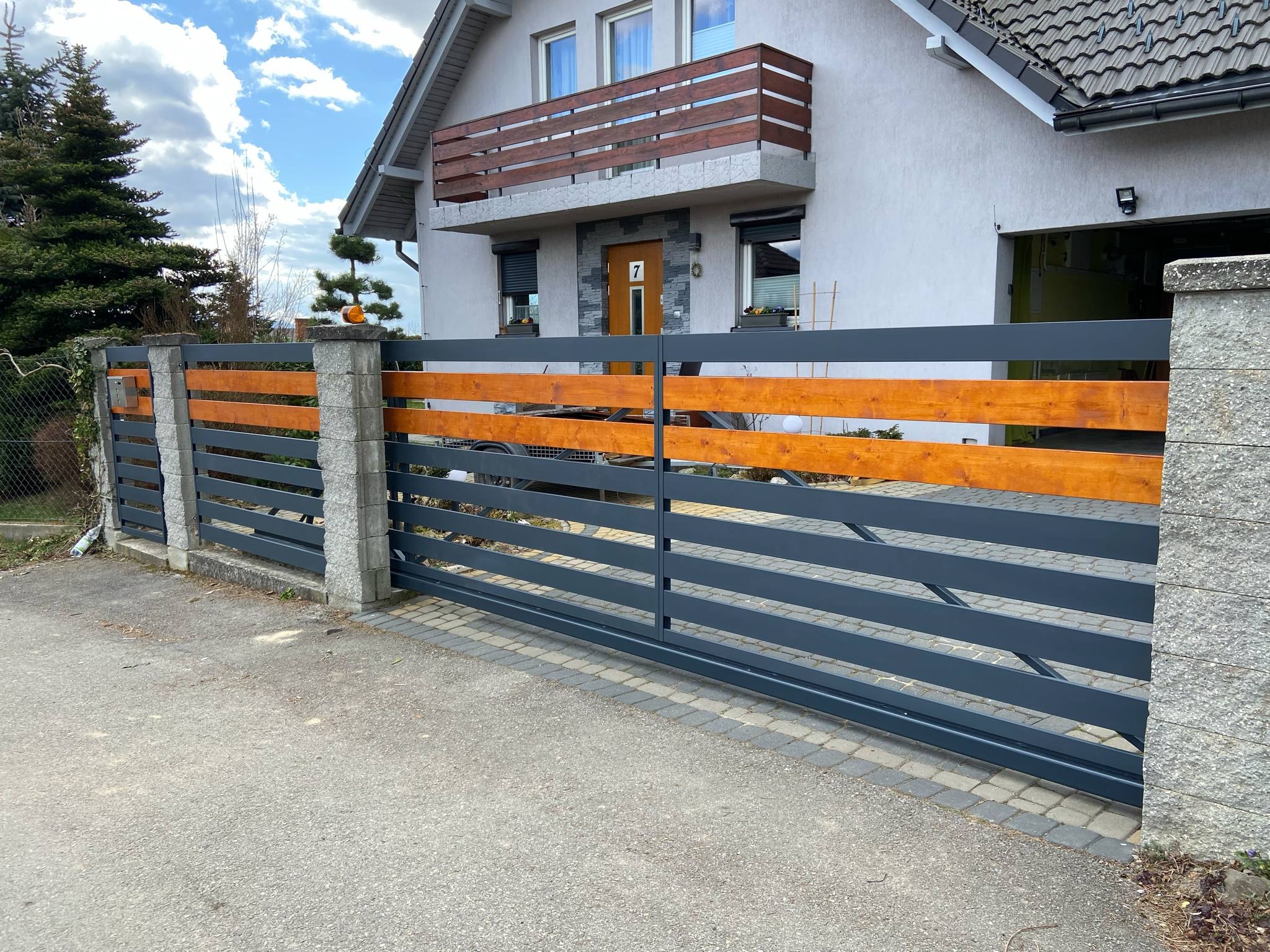 Nowoczesna, przesuwna brama wjazdowa z szarego metalu i drewnianymi akcentami, zamontowana przed domem z balkonem. Widoczne kamienne słupki i kostka brukowa.