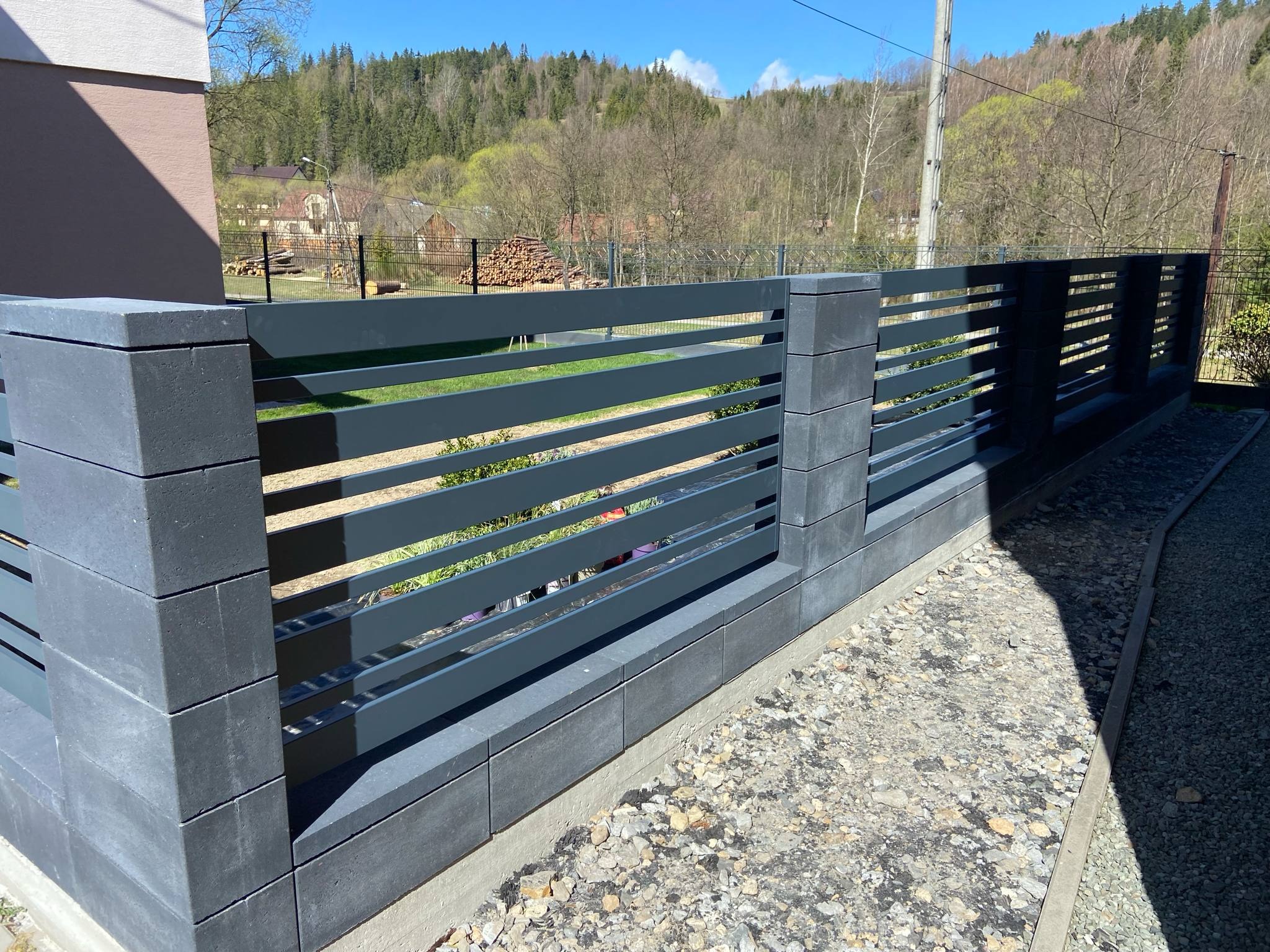 Nowoczesne ogrodzenie z poziomych, szarych paneli i betonowych słupków na tle zieleni i błękitnego nieba. Widok z bliska na fragment ogrodzenia.