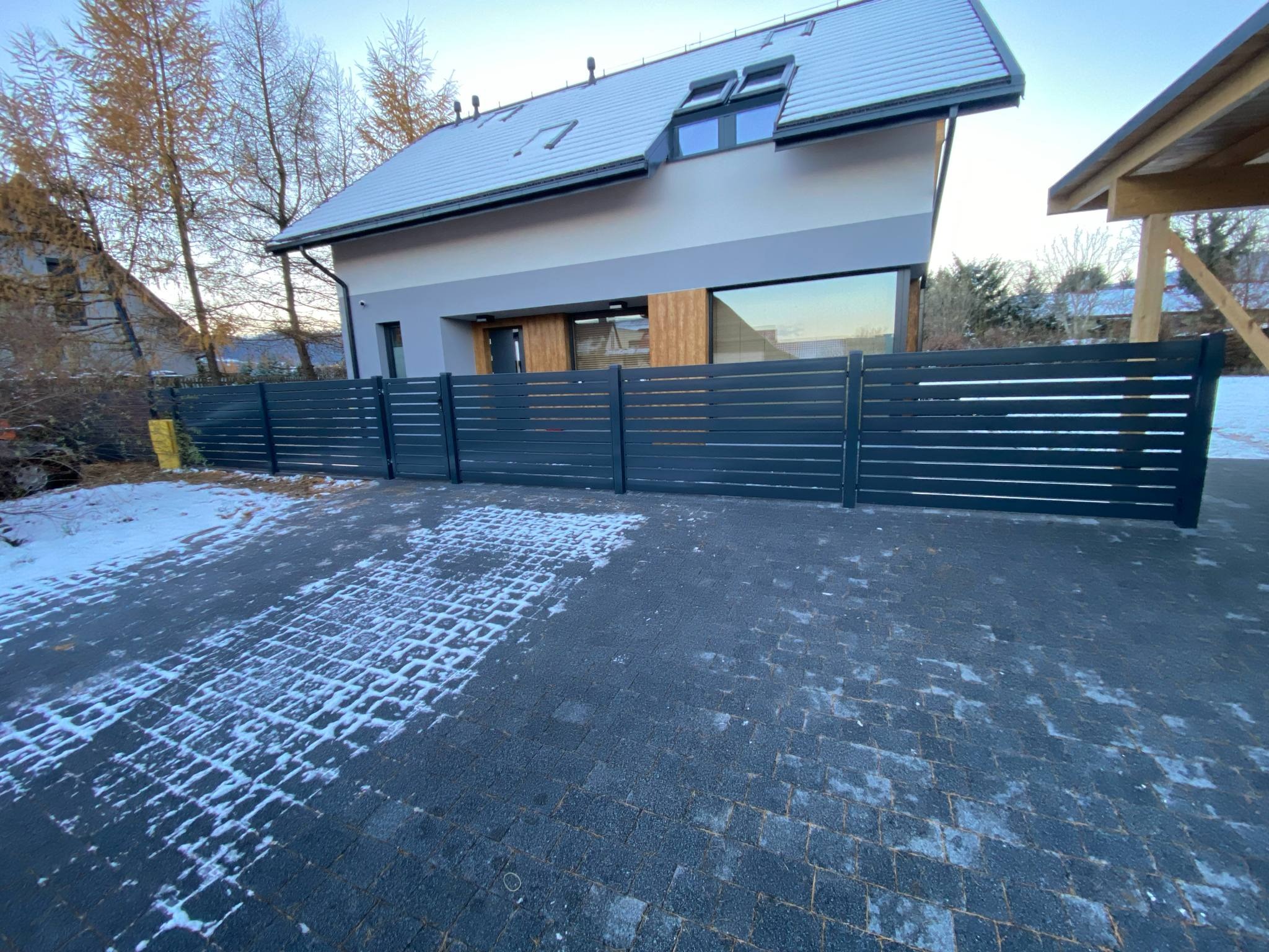 Nowoczesne, poziome ogrodzenie panelowe w kolorze antracytowym przed domem z szarym dachem, w zimowej scenerii. Widoczna kostka brukowa z resztkami śniegu.