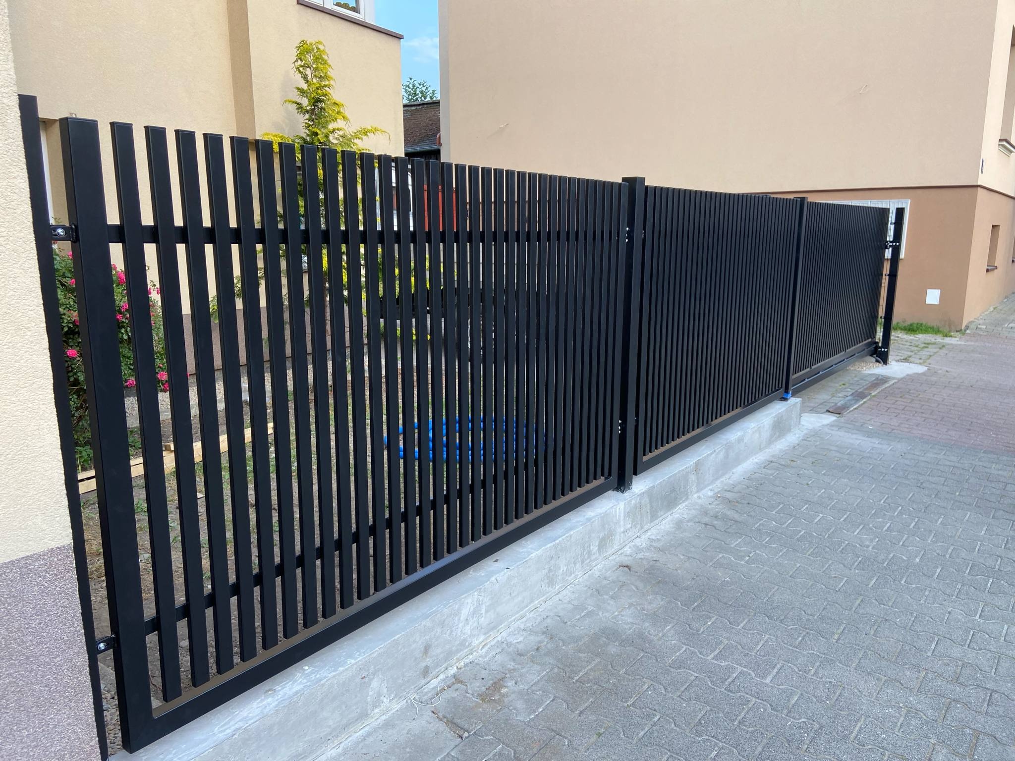 Nowoczesne, czarne ogrodzenie panelowe z pionowymi szczeblami, zamontowane na betonowej podmurówce, w tle budynek z beżową elewacją i zielenią.