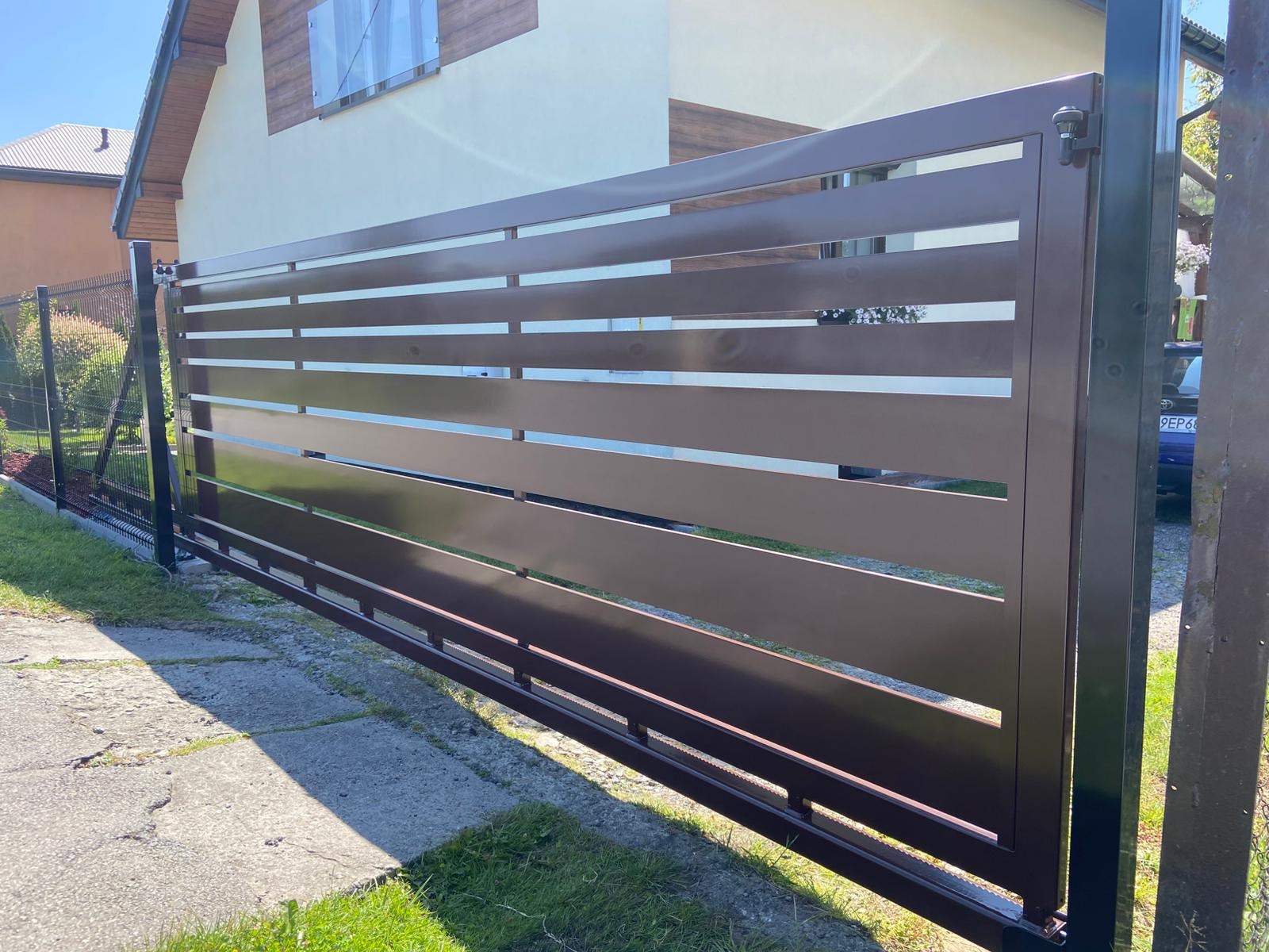 Nowoczesna, brązowa brama przesuwna z poziomymi panelami na tle domu z jasną elewacją i brązowym wykończeniem. Widoczne ogrodzenie panelowe po lewej stronie.