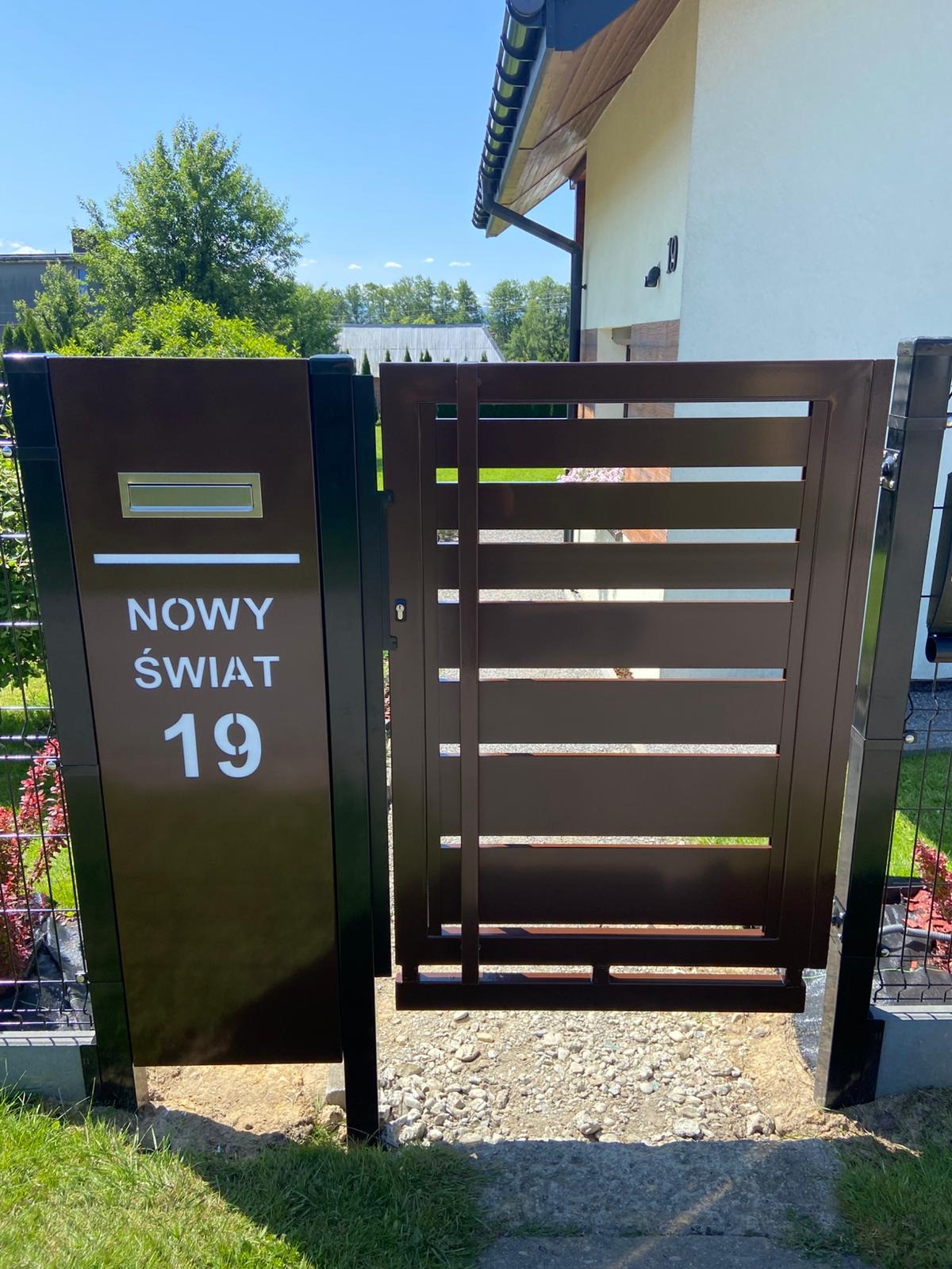 Nowoczesna, brązowa furtka z poziomymi szczeblami i skrzynką na listy z adresem 'Nowy Świat 19', zamontowana przy ogrodzeniu panelowym.