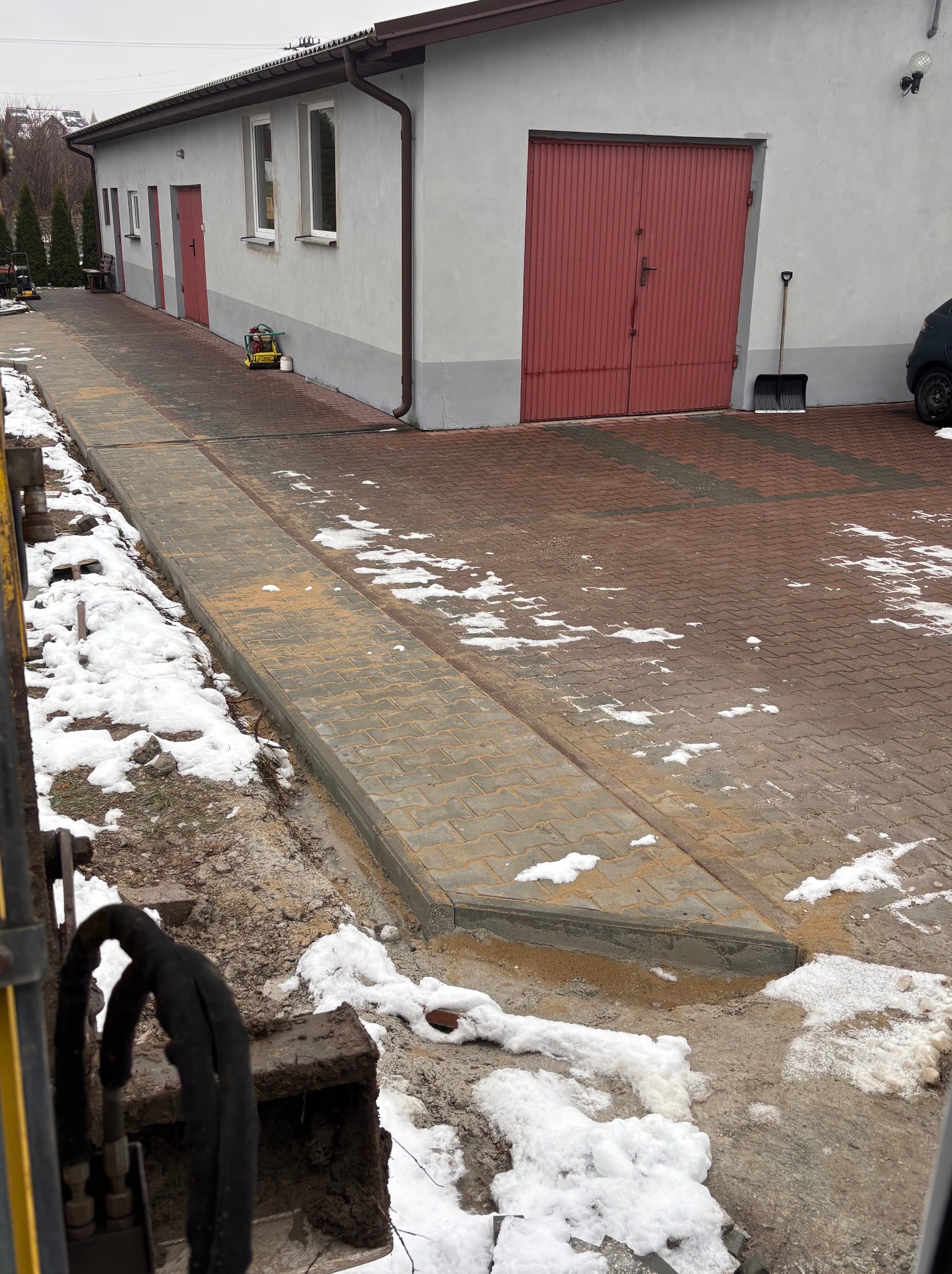 Ułożona kostka brukowa z obrzeżami, częściowo pokryta śniegiem, widoczny fragment koparki i budynek z czerwonymi drzwiami garażowymi w tle.