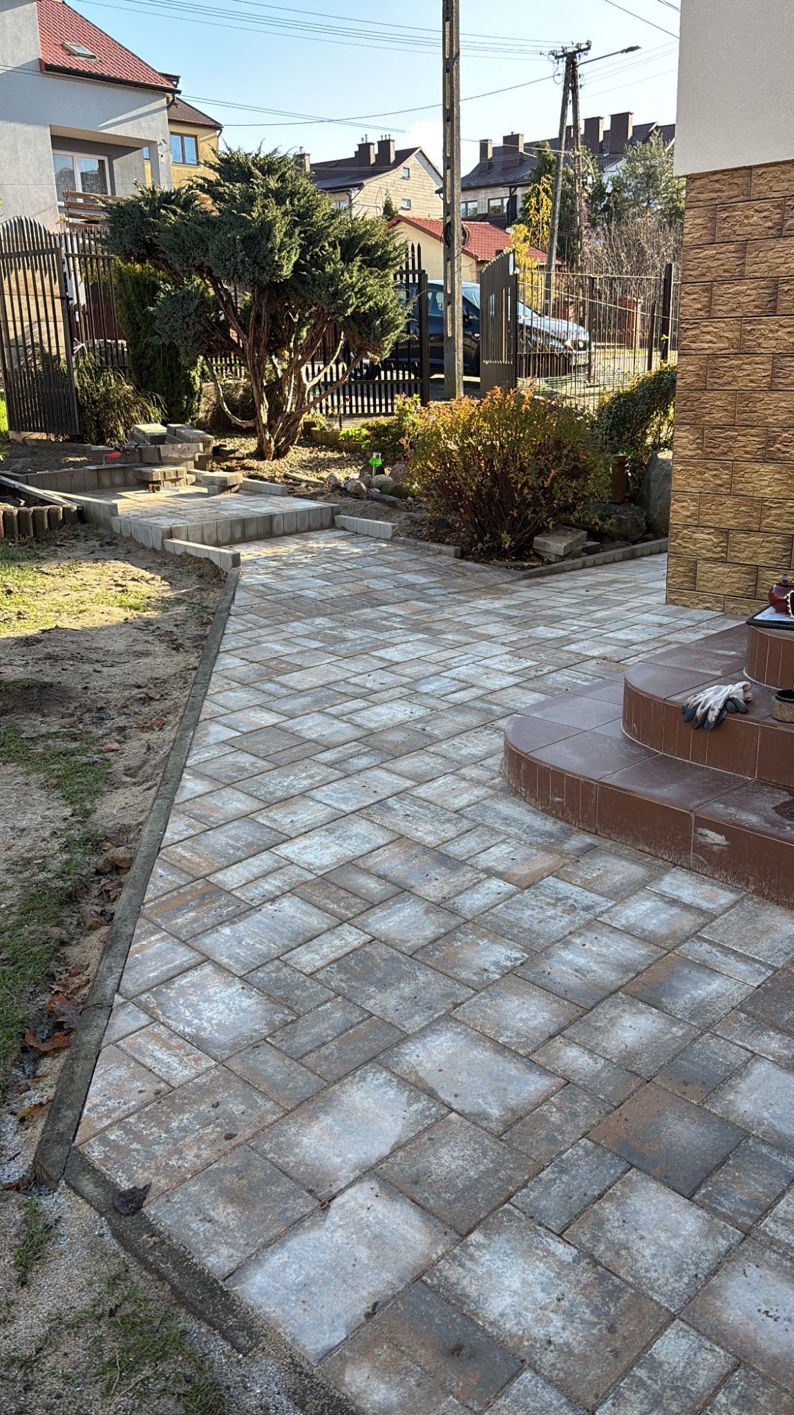 Ułożona kostka brukowa tworząca ścieżkę w ogrodzie, prowadząca do schodów wejściowych. Widoczne rękawice robocze na schodach i elementy zieleni w tle.