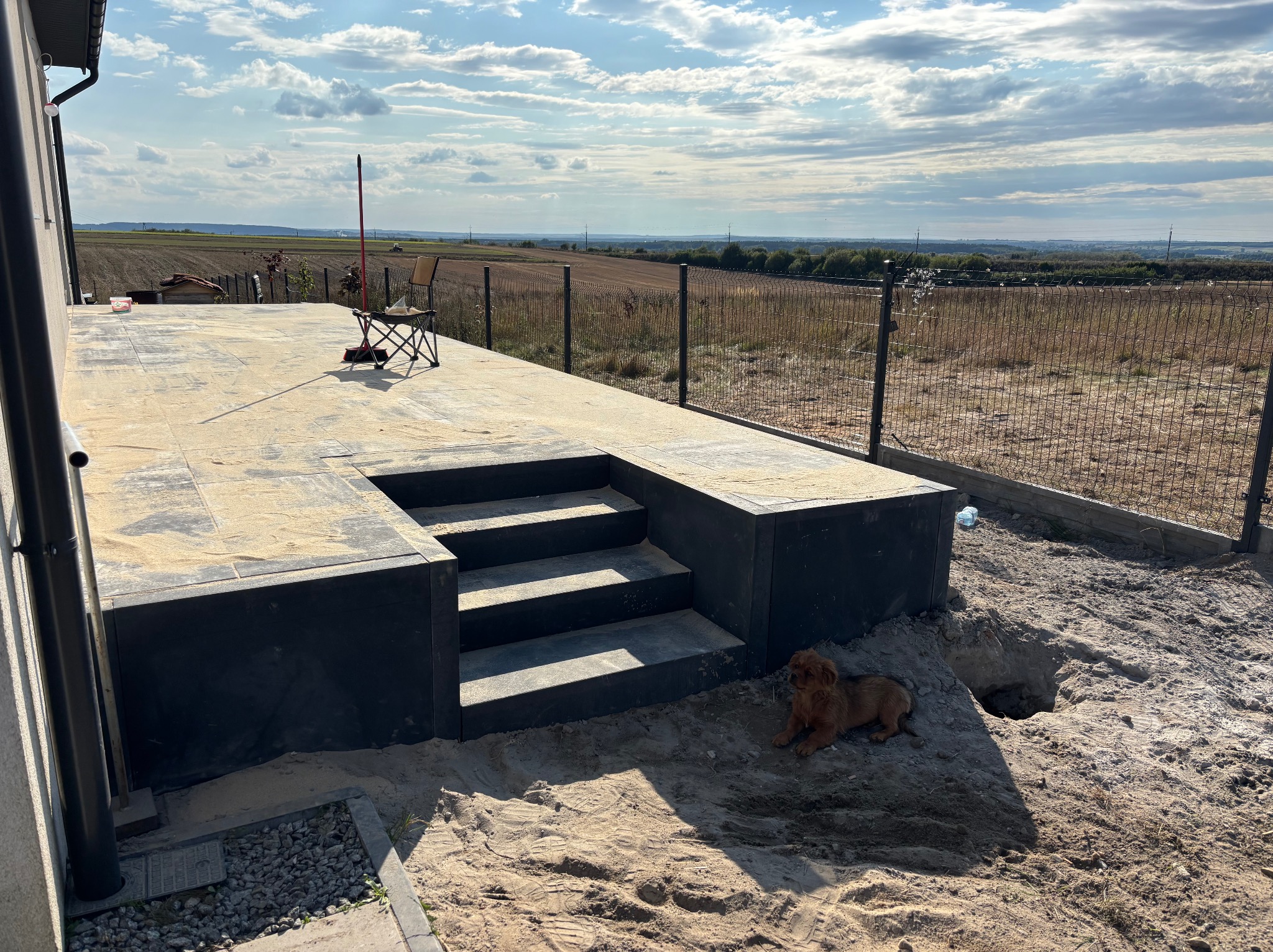 Wykończony taras z czarnej kostki z schodami, otoczony piaskiem i nowym ogrodzeniem z siatki. W tle rozległe pole i błękitne niebo. Pies leży obok wykopanego dołu.