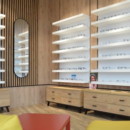 Studio 77 - Nowoczesny salon optyczny: drewniane panele, białe półki z okularami, owalne lustro i komody. Jasne oświetlenie, minimalistyczny design i żółte akcenty kolorystyczne.
