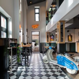 Studio 77 - Wnętrze restauracji w stylu retro: biało-czarna podłoga w szachownicę, błękitny skuter, bar z drewnianym blatem, plakaty vintage i wysokie okna wpadające do środka.