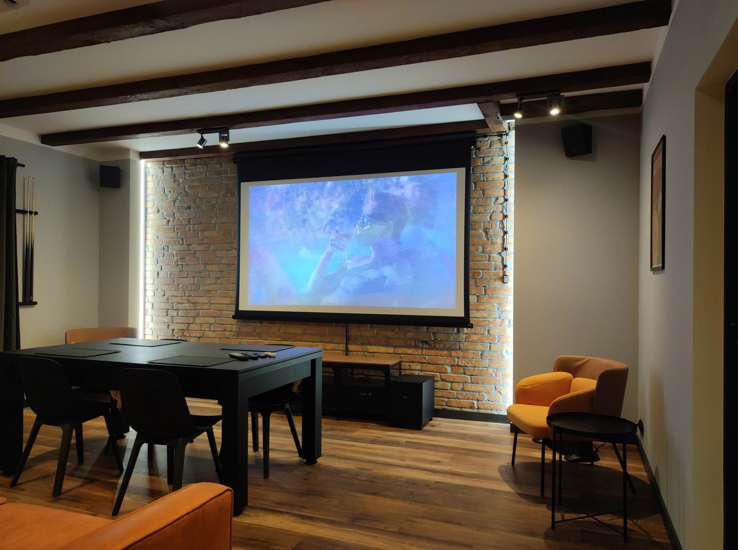 Kącik kinowy w salonie: projektor z ekranem na ceglanej ścianie, czarny stół z krzesłami i pomarańczowy fotel. Drewniane belki sufitowe i punktowe oświetlenie tworzą przytulny klimat.
