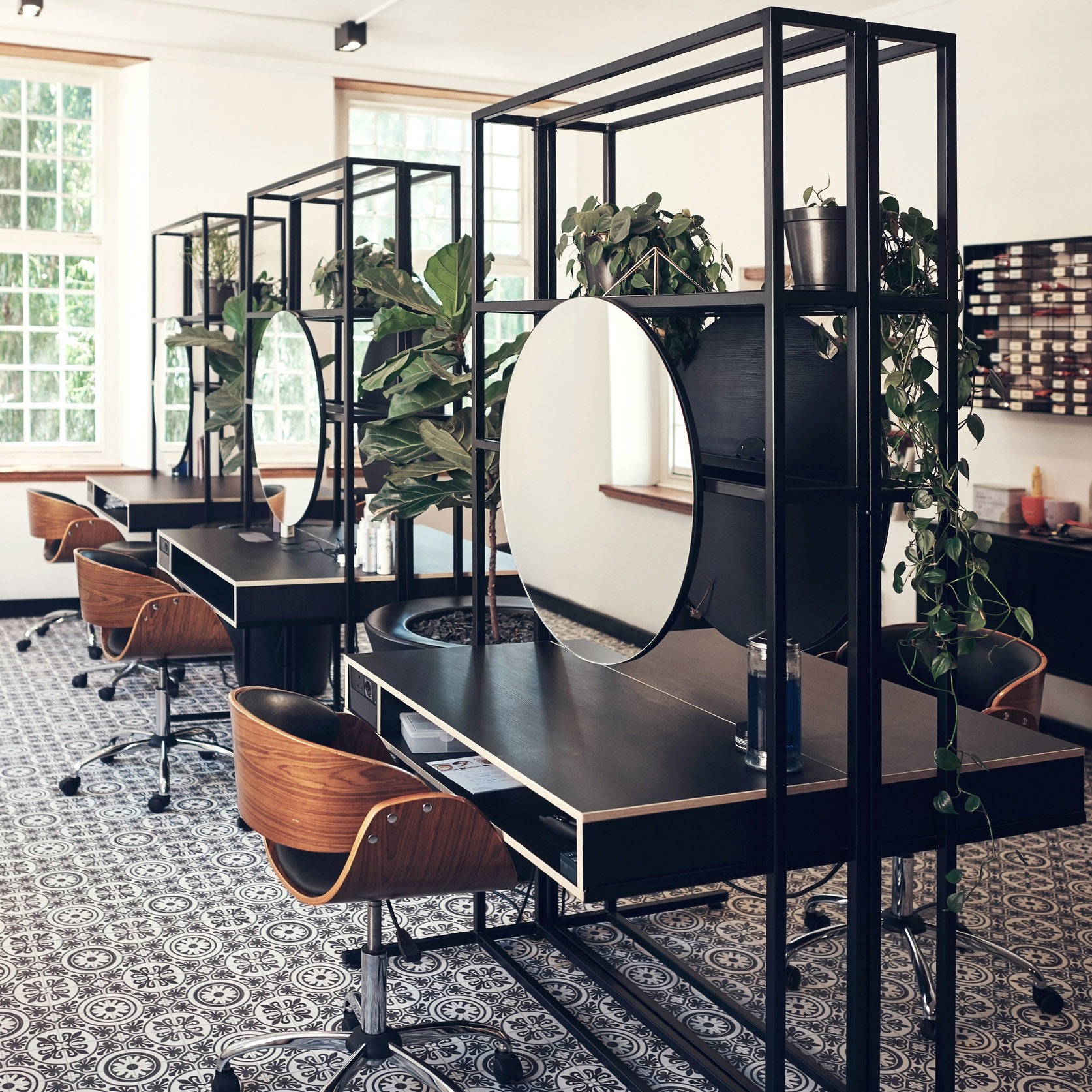 Nowoczesne stanowiska fryzjerskie z okrągłymi lustrami, roślinami doniczkowymi i drewnianymi krzesłami na tle wzorzystej podłogi w jasnym salonie.