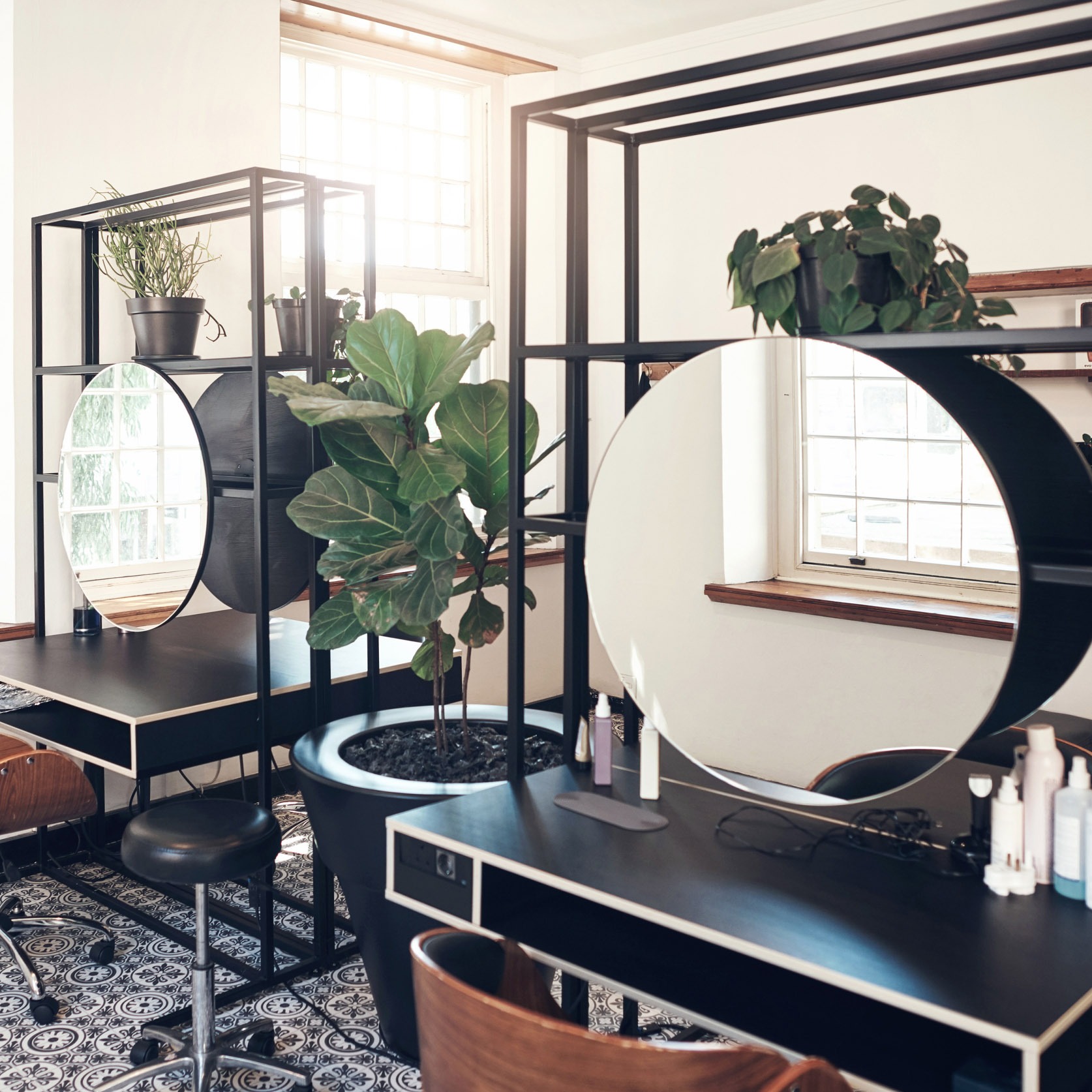 Nowoczesne stanowisko fryzjerskie z okrągłym lustrem, czarnym blatem i roślinami doniczkowymi. Minimalistyczny design, jasne wnętrze, wzorzysta podłoga.