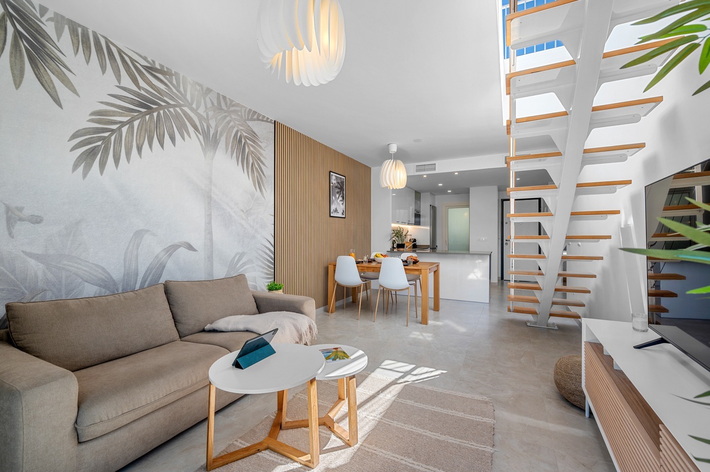 Jasne wnętrze mieszkania z motywem tropikalnym na ścianie, drewnianymi panelami i schodami. Widoczny salon z sofą, stolikami i jadalnia z kuchnią w tle.
