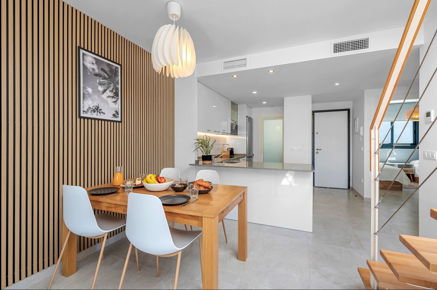 Jasna jadalnia z drewnianym stołem, białymi krzesłami i ścianą z drewnianych paneli. W tle widoczna kuchnia i schody z drewnianymi stopniami.