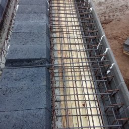 ErbudDom Nicoll Mikołajczyk - Zbrojenie fundamentów z bloczków betonowych, połączonych białą pianką montażową. Widoczne drewniane elementy szalunku i wystająca poza kadr stal zbrojeniowa.