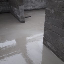 ErbudDom Nicoll Mikołajczyk - Świeżo wylana posadzka samopoziomująca w surowym wnętrzu z szarymi ścianami z bloczków. Widoczne refleksy światła na gładkiej powierzchni.