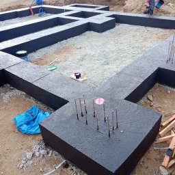 ErbudDom Nicoll Mikołajczyk - Czarne, zabezpieczone fundamenty domu w trakcie budowy, z wystającymi prętami zbrojeniowymi. W tle widoczny pracownik z wiadrem i wykop pod fundamenty.