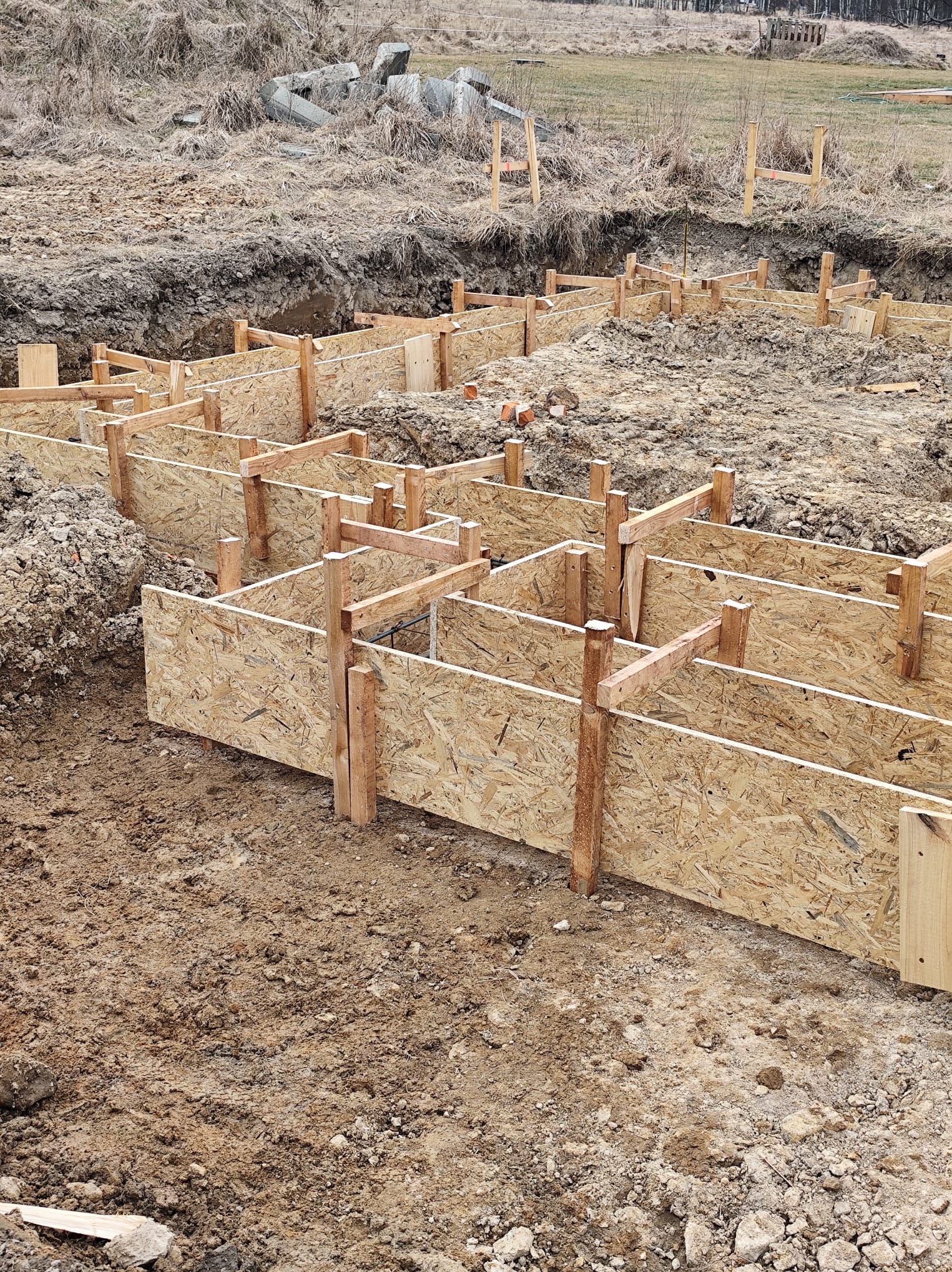 Szalunek z płyty OSB i drewnianych stempli, przygotowany do wylania fundamentów pod budowę domu. Widok z góry na plac budowy w pochmurny dzień.