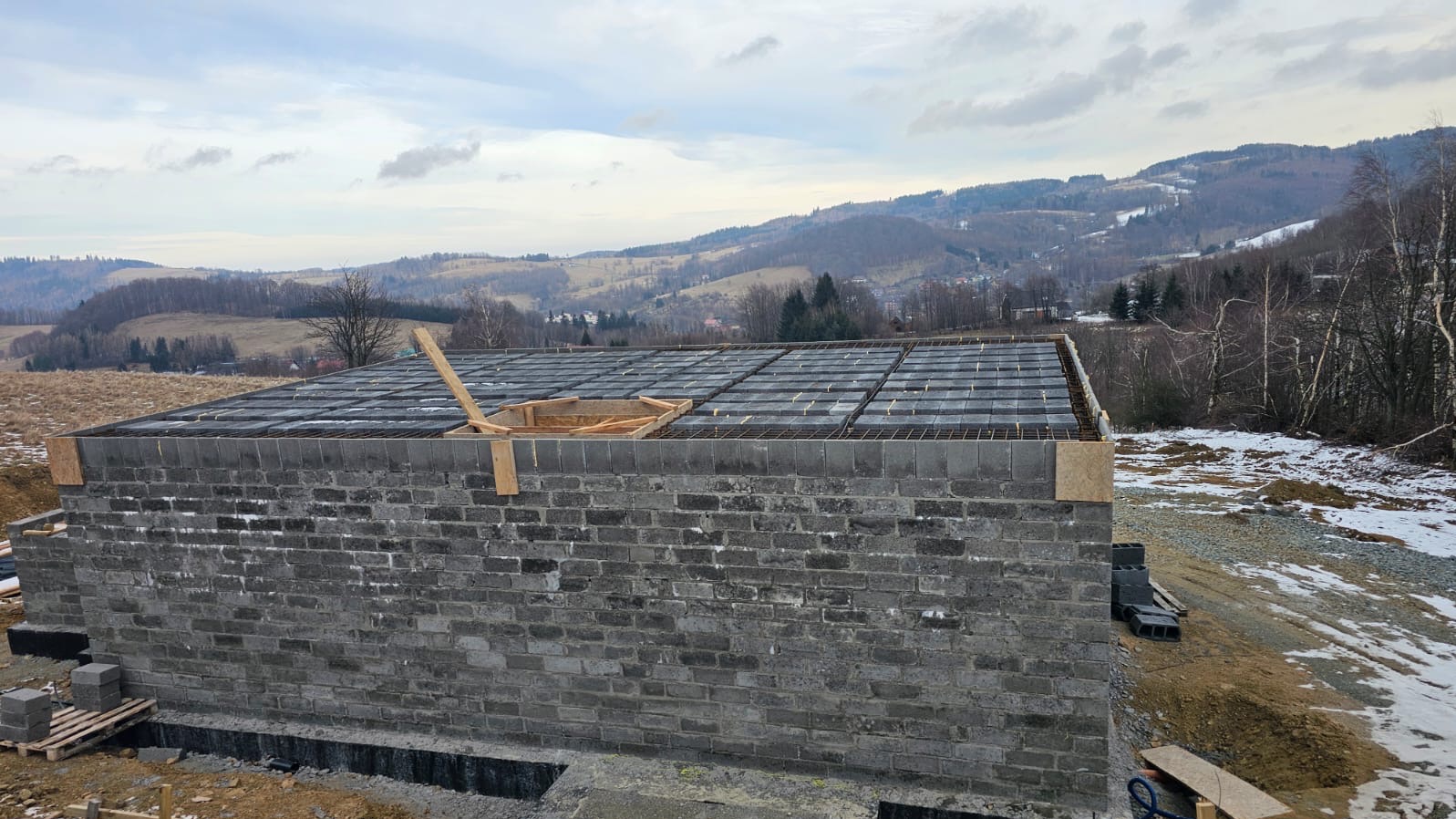 Murowany budynek w trakcie budowy, z widoczną konstrukcją stropu i zbrojeniami. W tle pagórkowaty krajobraz i resztki śniegu.