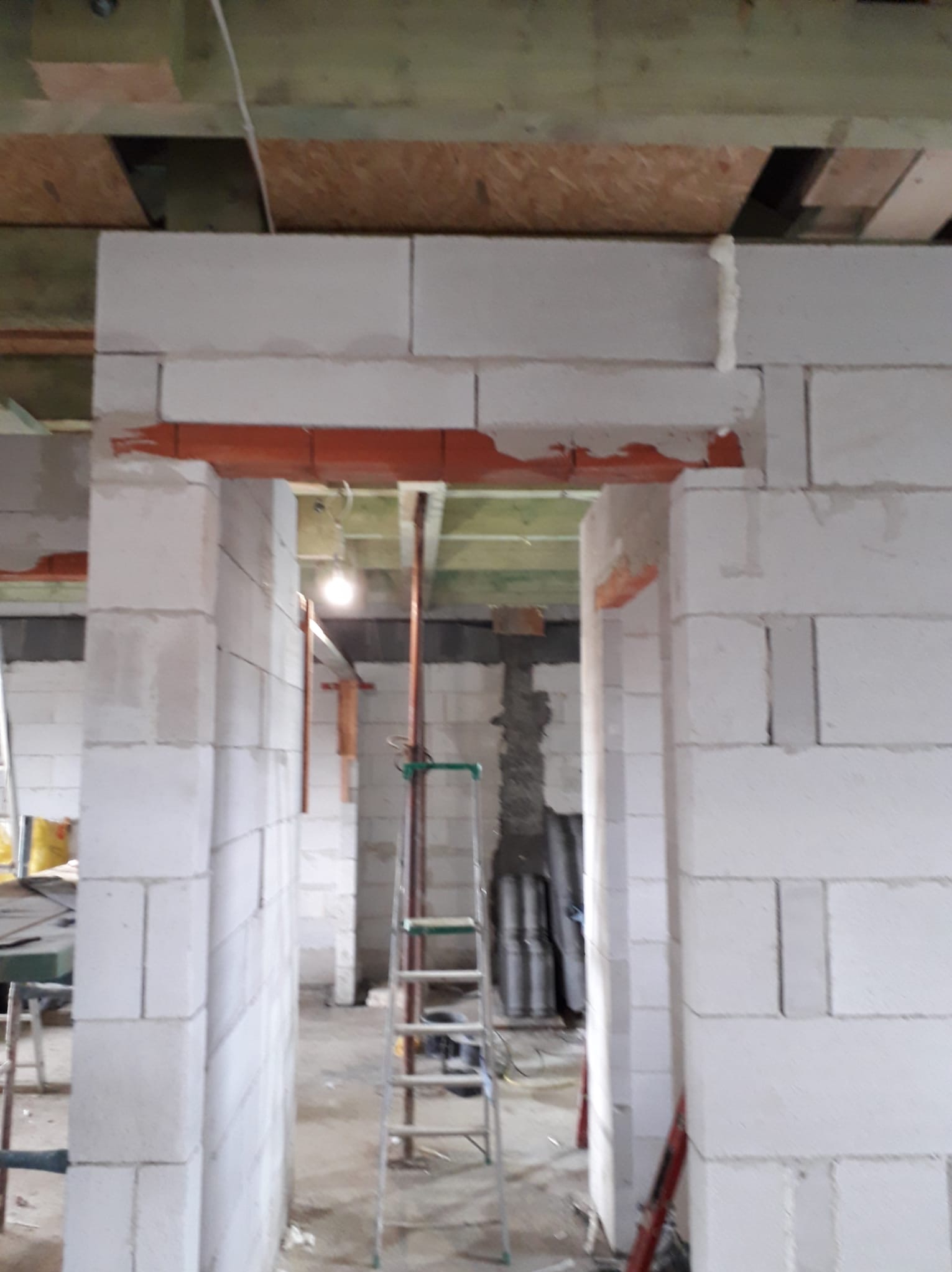 Wnętrze w trakcie budowy: białe bloczki ścian działowych, widoczne nadproże z cegły, drabina. Surowy stan, widoczne elementy konstrukcji stropu.
