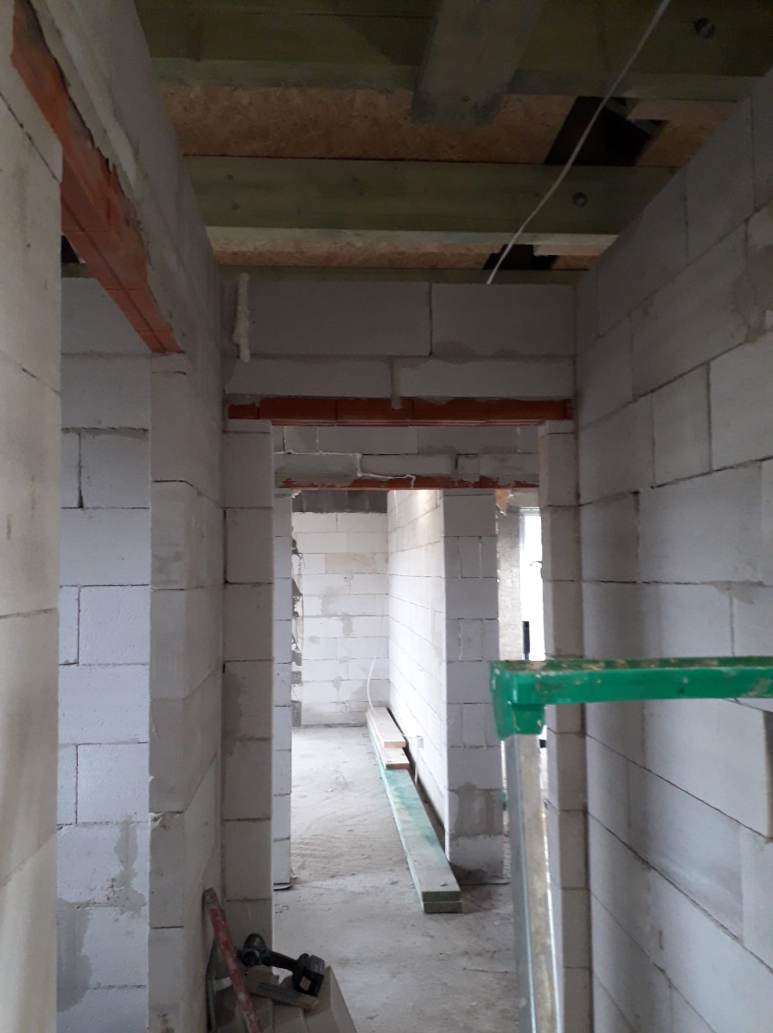 Wnętrze w trakcie budowy: białe ściany z betonu komórkowego, drewniane belki stropowe, widoczne otwory drzwiowe i okienne, elementy rusztowania. Surowy stan wykończenia.