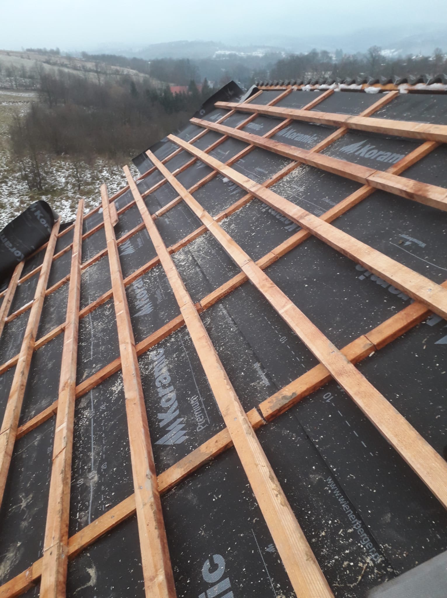 Konstrukcja dachu z łatami drewnianymi na czarnej membranie, widok z góry. Fragment pokrycia dachowego z elementami mocującymi. Tło: zalesione wzgórza w zimowej scenerii.