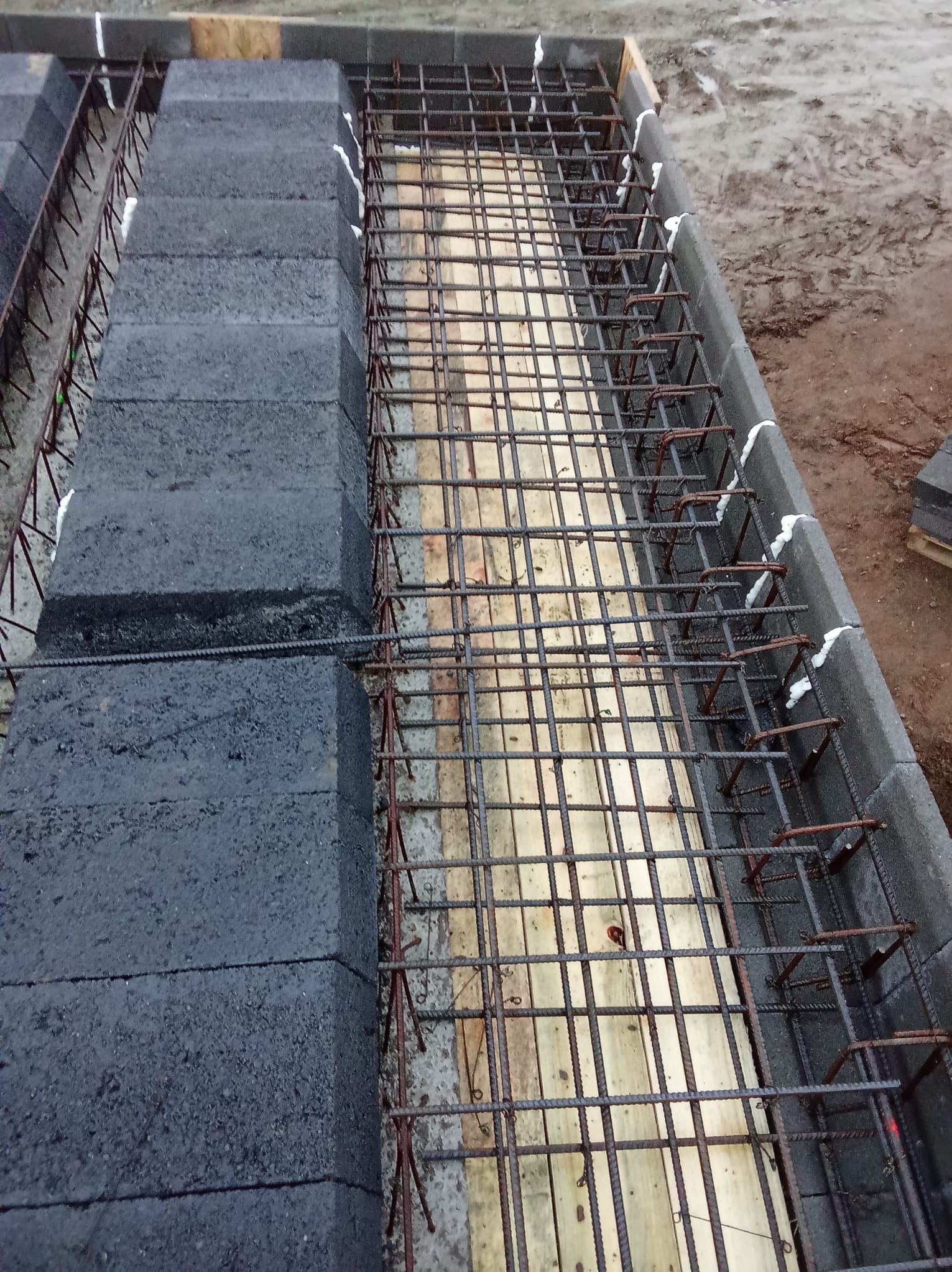 Zbrojenie fundamentów z bloczków betonowych, połączonych białą pianką montażową. Widoczne drewniane elementy szalunku i wystająca poza kadr stal zbrojeniowa.