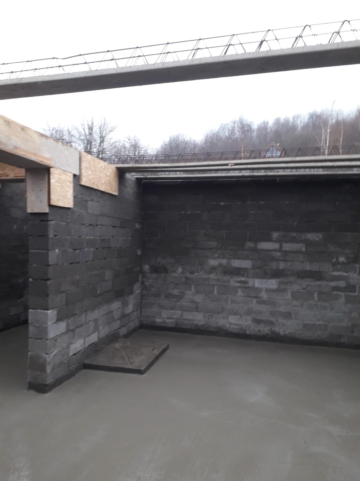 Surowy stan: Ściany z szarej cegły, świeżo wylana betonowa podłoga, drewniane elementy konstrukcyjne. Widok na fragment budynku w trakcie budowy.