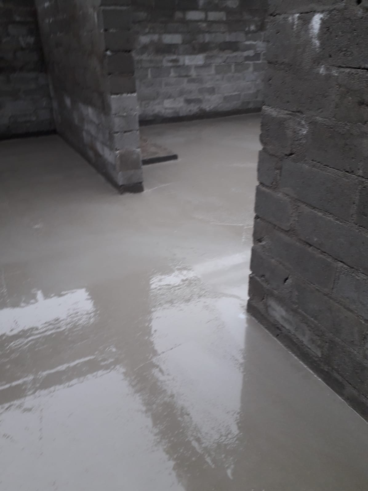 Świeżo wylana posadzka samopoziomująca w surowym wnętrzu z szarymi ścianami z bloczków. Widoczne refleksy światła na gładkiej powierzchni.