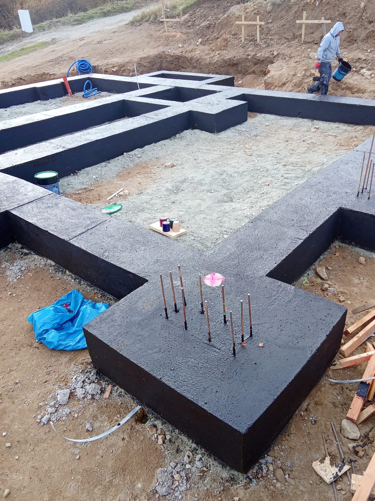 Czarne, zabezpieczone fundamenty domu w trakcie budowy, z wystającymi prętami zbrojeniowymi. W tle widoczny pracownik z wiadrem i wykop pod fundamenty.
