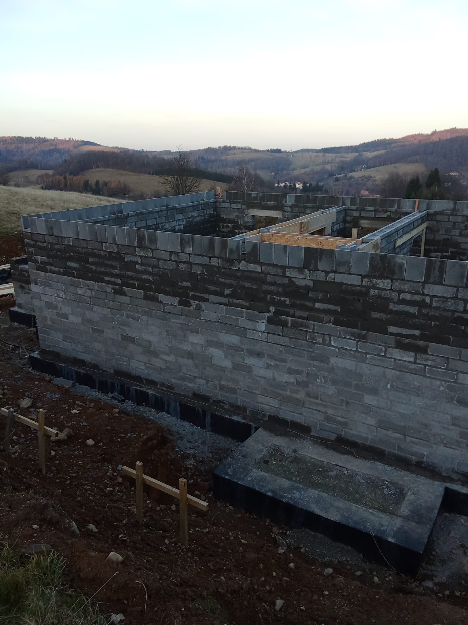 Wznoszenie ścian z bloczków betonowych na fundamencie z izolacją, widok na pagórkowaty krajobraz w tle. Etap budowy domu z surowych materiałów.