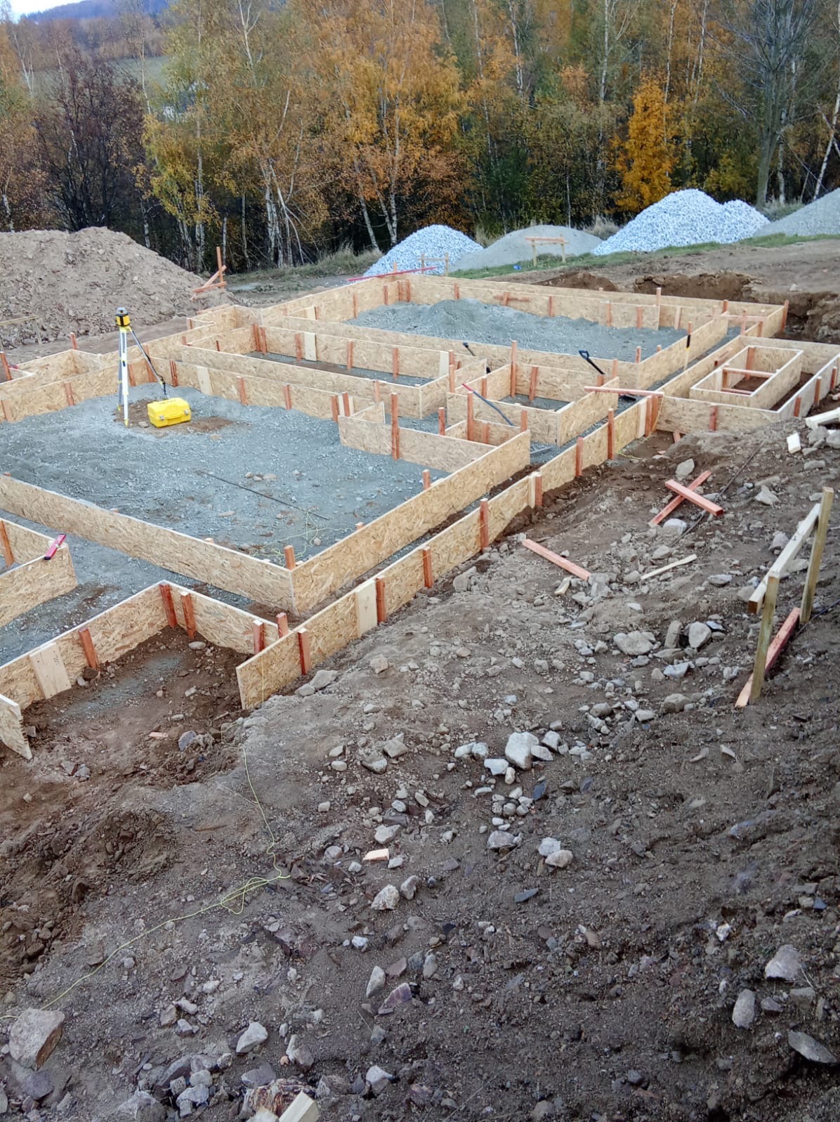 Szalunek pod fundamenty domu jednorodzinnego, wypełniony żwirem. W tle hałdy żwiru i jesienny krajobraz. Widoczny niwelator laserowy.