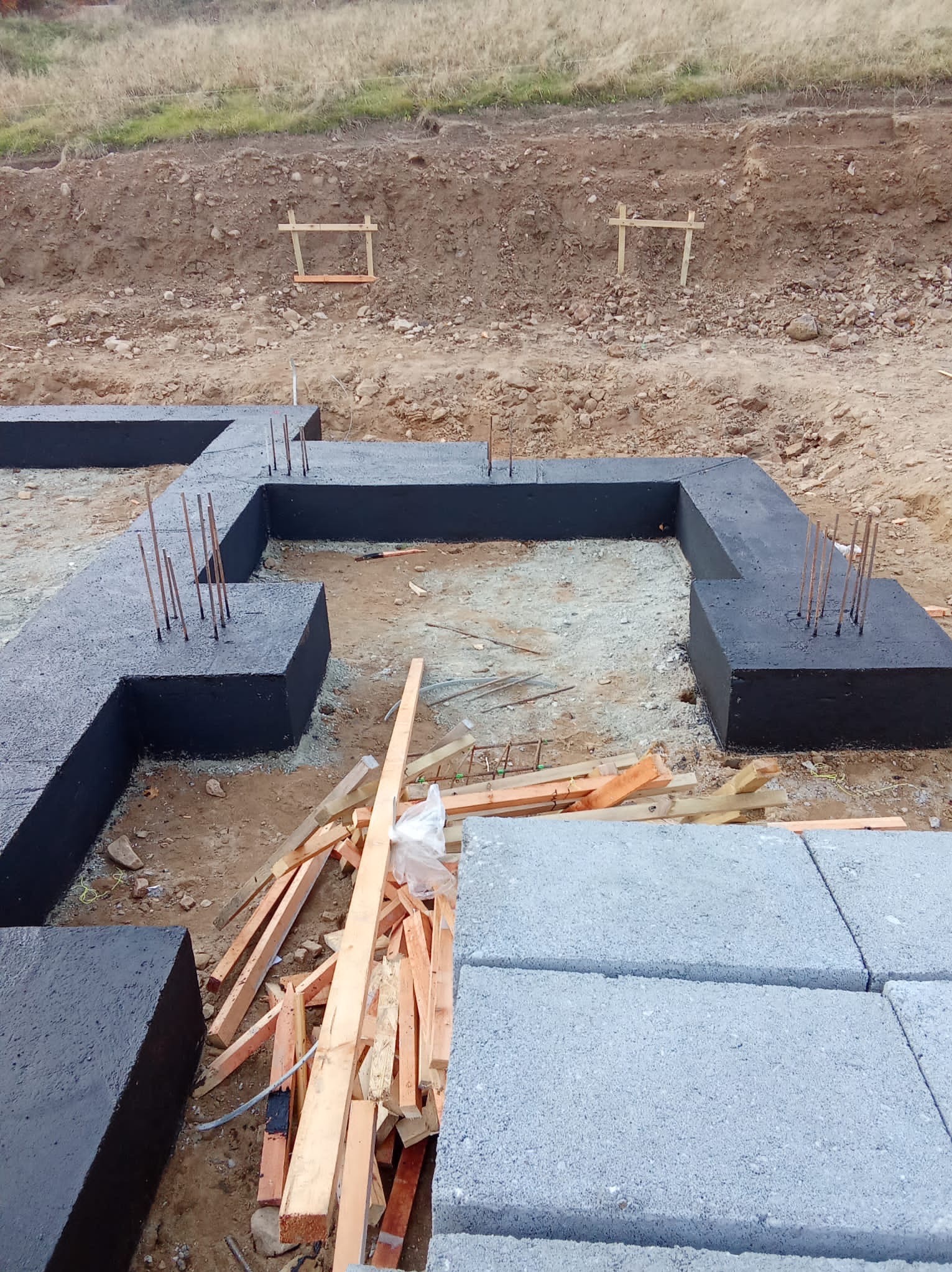 Zabezpieczone, czarne fundamenty domu z wystającymi prętami zbrojeniowymi, ułożone na ziemi. W tle wykop i drewniane elementy szalunku.