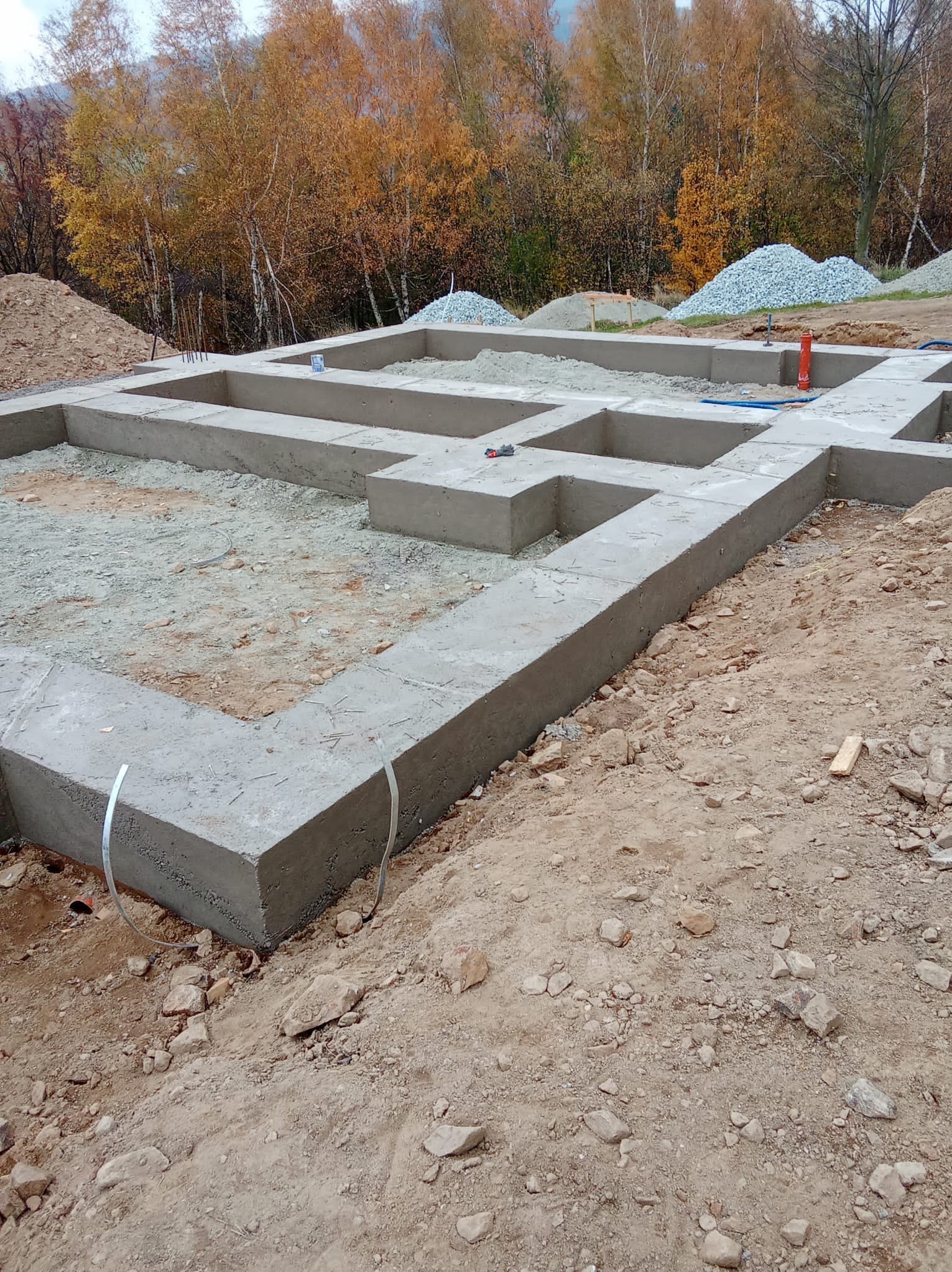 Wylane fundamenty domu jednorodzinnego na tle jesiennego lasu. Widoczne zbrojenia, instalacje i hałdy żwiru. Kąt ujęcia z dołu, podkreślający solidność konstrukcji.