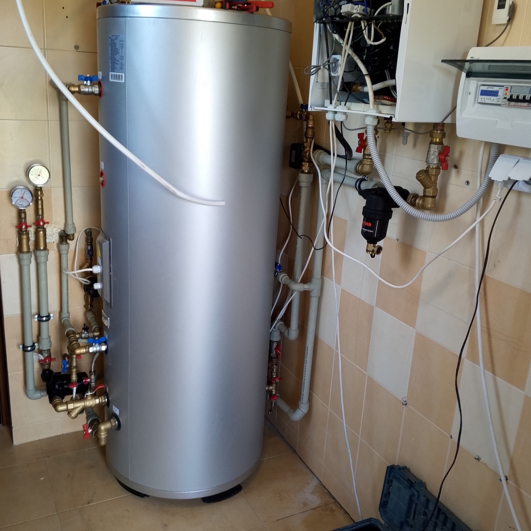 Srebrny zasobnik ciepłej wody użytkowej z podłączonymi rurami miedzianymi i plastikowymi, manometry, filtry oraz skrzynka elektryczna na ścianie wyłożonej kafelkami.