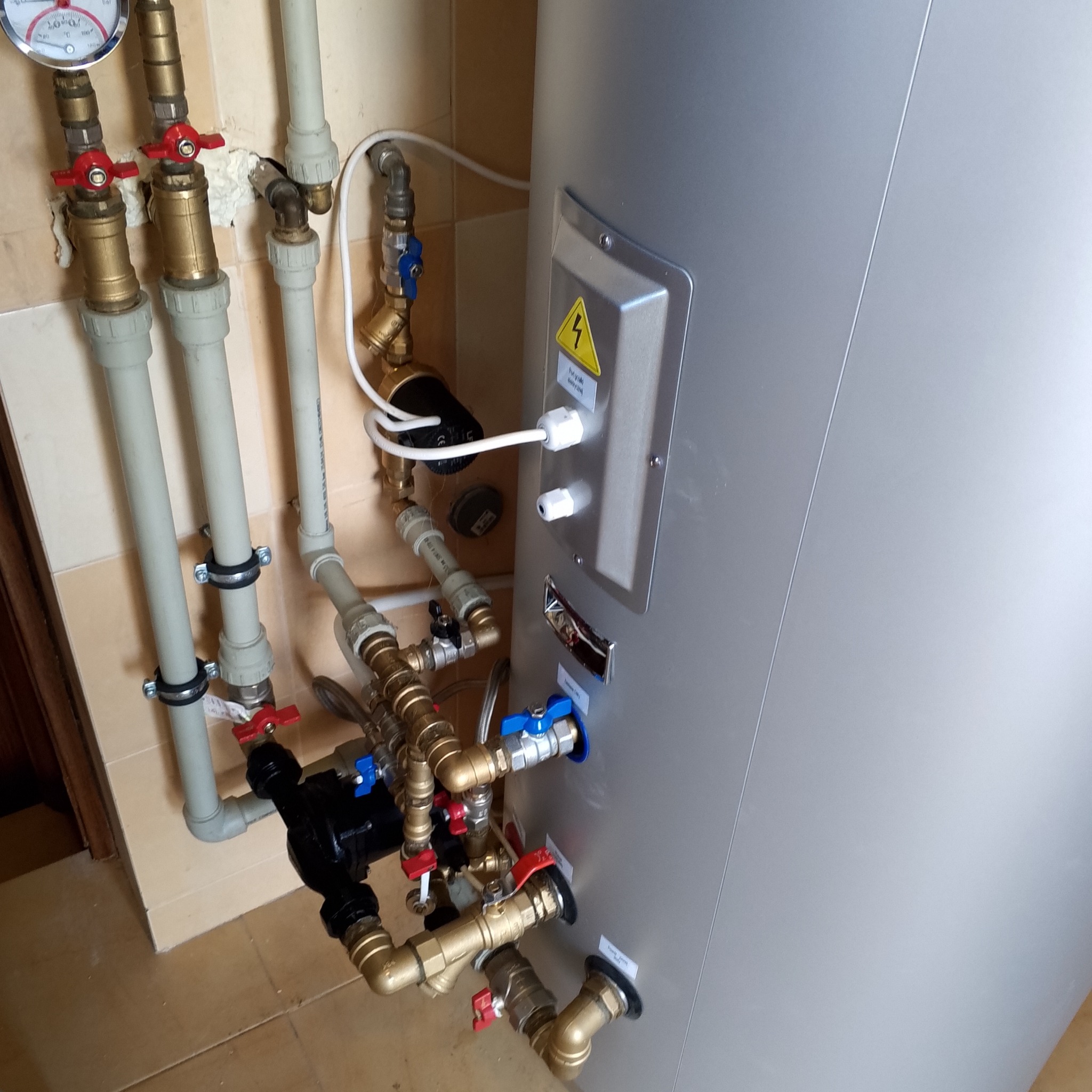 Szary zasobnik ciepłej wody użytkowej z podłączonymi rurami miedzianymi i plastikowymi, zaworami z czerwonymi i niebieskimi uchwytami oraz manometrem, zamontowany w pomieszczeniu z beżowymi płytkami.