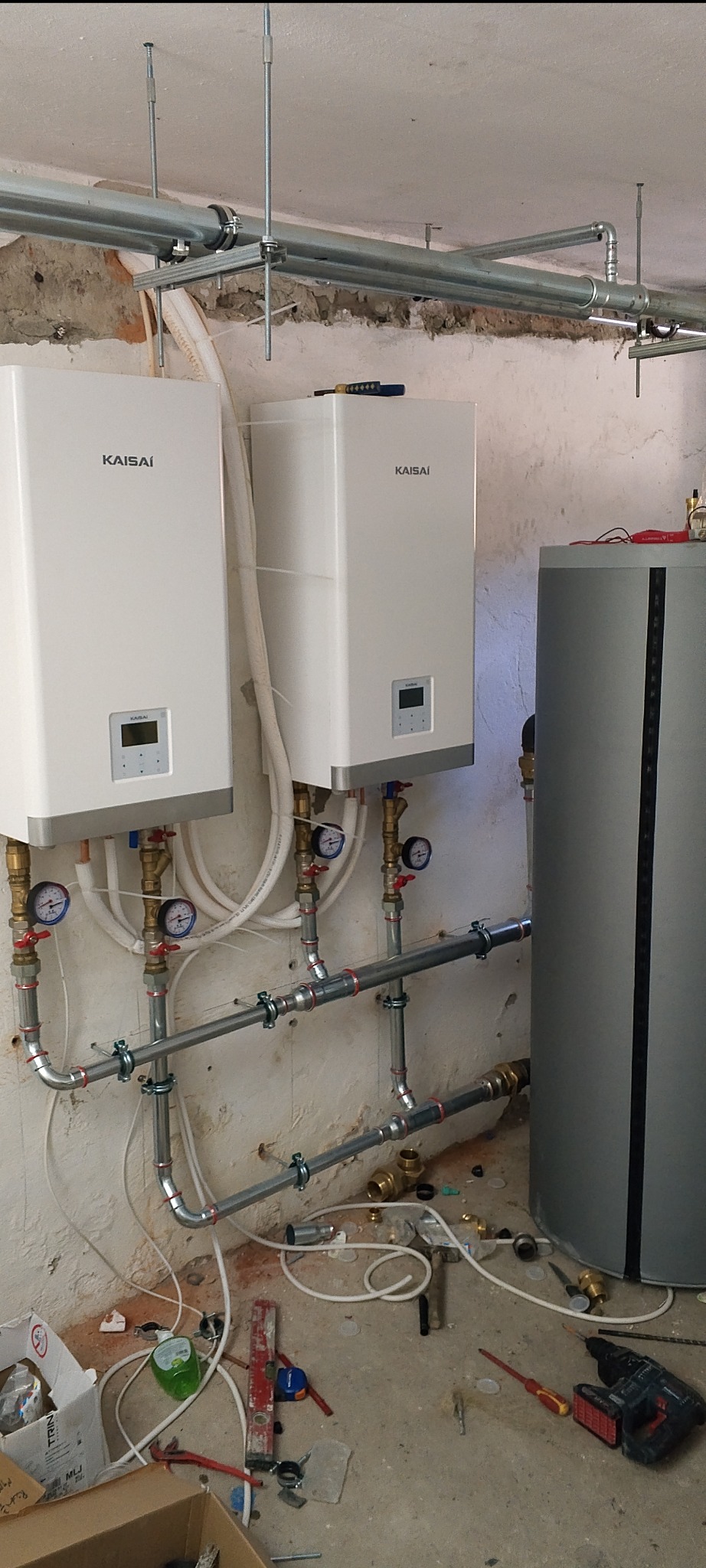 Dwa białe, wiszące piece gazowe marki Kaisai z podłączonymi rurami i manometrami, oraz szary zasobnik na wodę, zamontowane na tle surowej ściany z widocznymi instalacjami. Na podłodze widoczne...