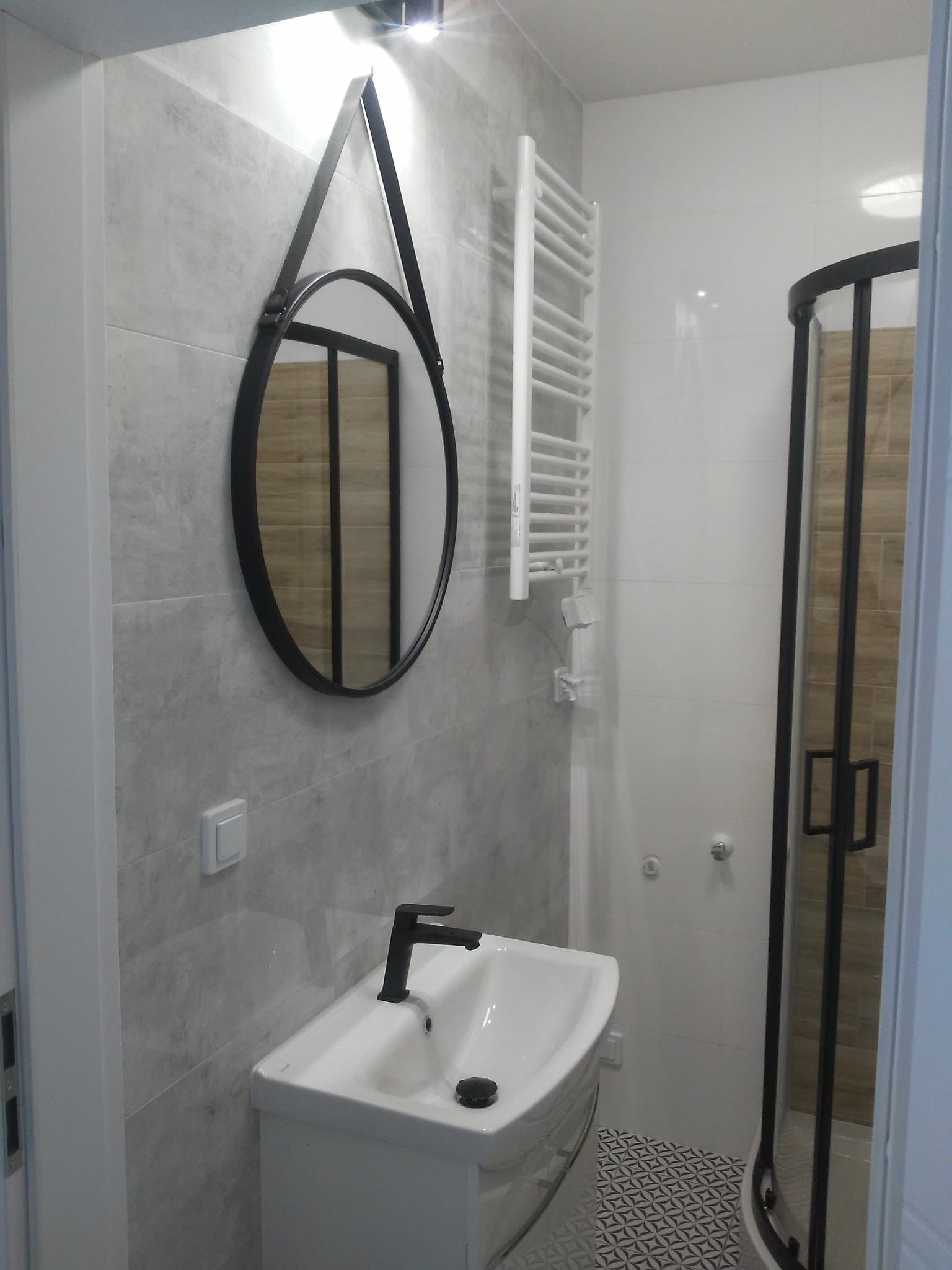 Nowoczesna łazienka z owalnym lustrem w czarnej ramie, białą umywalką z czarną armaturą, grzejnikiem drabinkowym i kabiną prysznicową z czarnymi profilami.