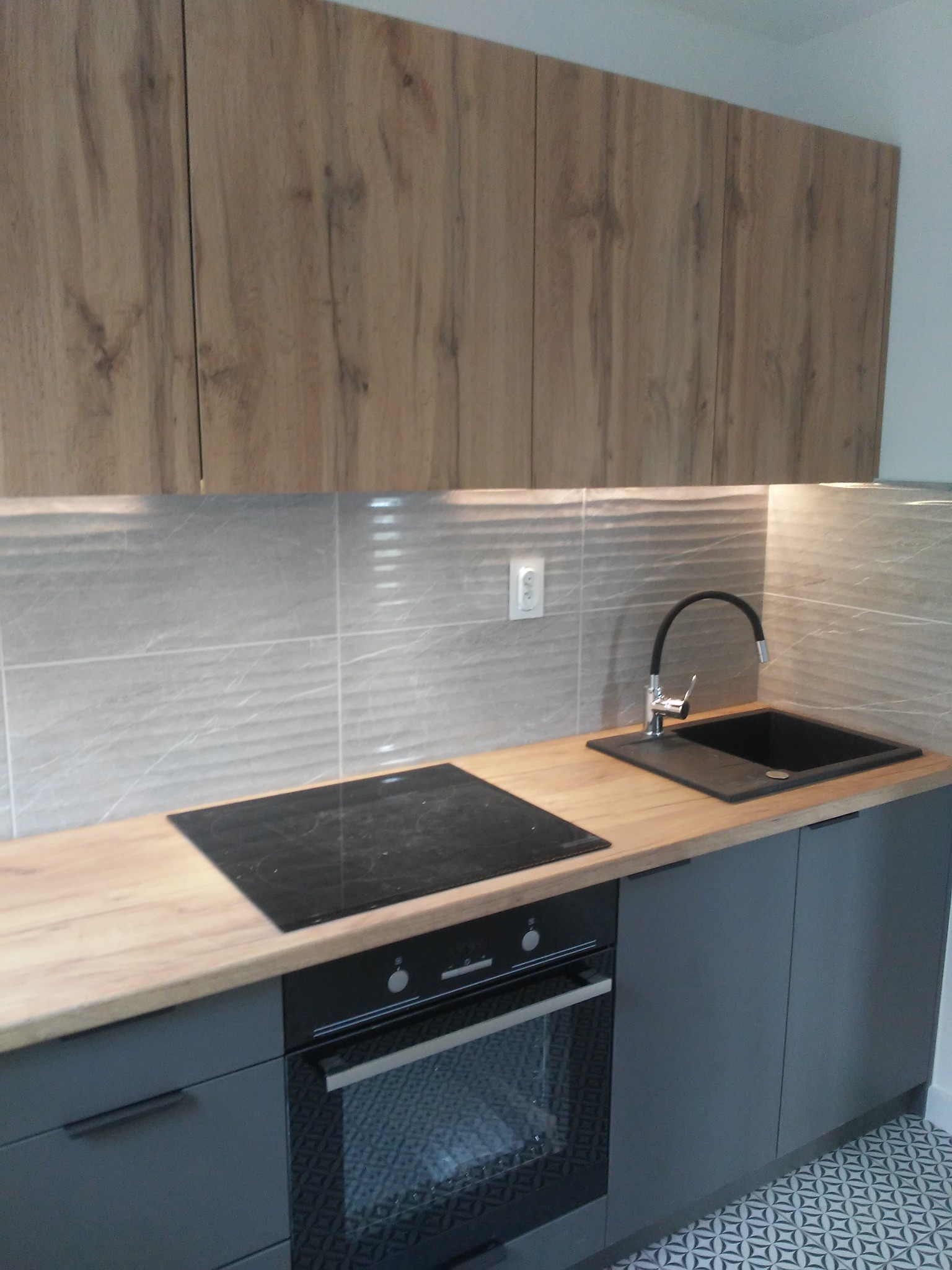 Nowoczesna kuchnia z drewnianymi szafkami wiszącymi i szarymi szafkami dolnymi. Na blacie płyta indukcyjna i czarny zlew. W tle szare płytki z delikatnym wzorem.