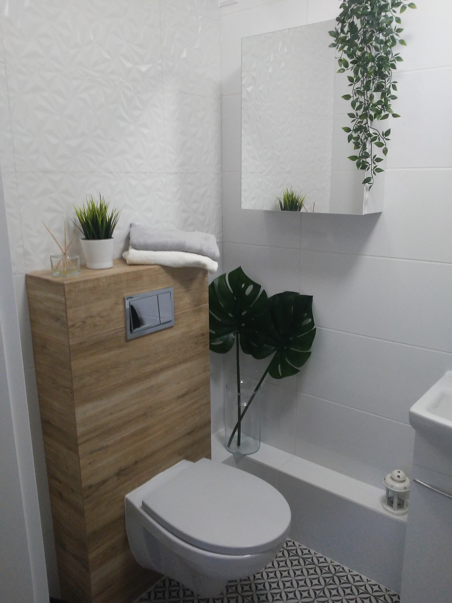 Nowoczesna toaleta w jasnych barwach z drewnianą obudową geberitu, dekoracyjnymi płytkami i roślinami. Minimalistyczny design z akcentami natury.