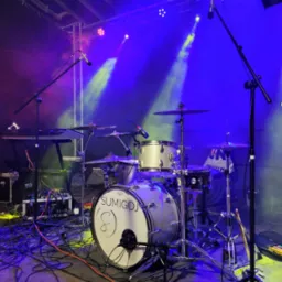 Perkusja zespołu Sumigoj na scenie w oświetleniu estradowym. Widoczne mikrofony, kable i klawisze. Dynamiczne światła w kolorach niebieskim i żółtym tworzą atmosferę koncertu.
