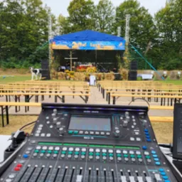 Konsoleta Allen&Heath SQ-5 na tle sceny plenerowej z dekoracjami dożynkowymi i rzędami ławek. Na lewo stoi krowa.