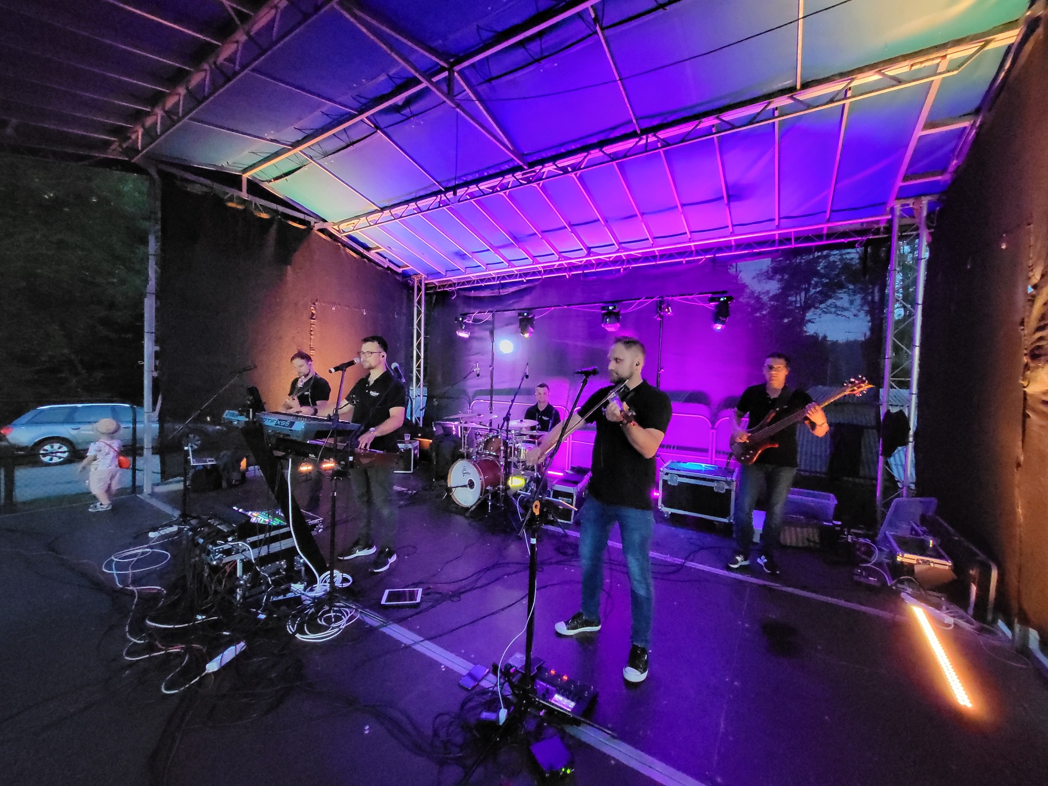 Zespół muzyczny gra na scenie oświetlonej fioletowym światłem. Widoczni muzycy z instrumentami: skrzypce, gitara basowa, keyboard, perkusja. Z przodu kable i sprzęt.