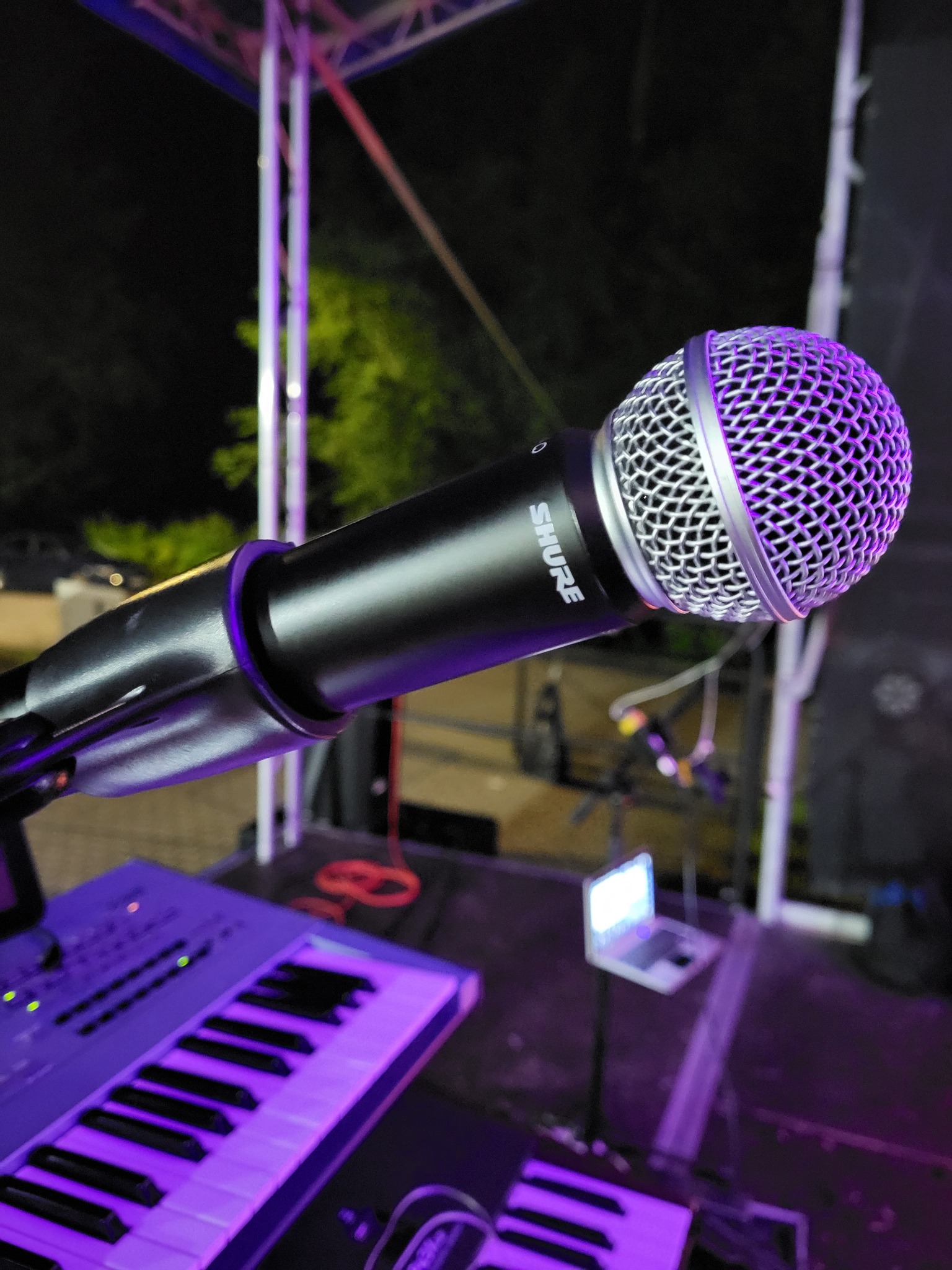 Mikrofon Shure na statywie, oświetlony fioletowym światłem, z klawiaturą i laptopem w tle, ustawiony na scenie. Widoczna konstrukcja sceniczna.