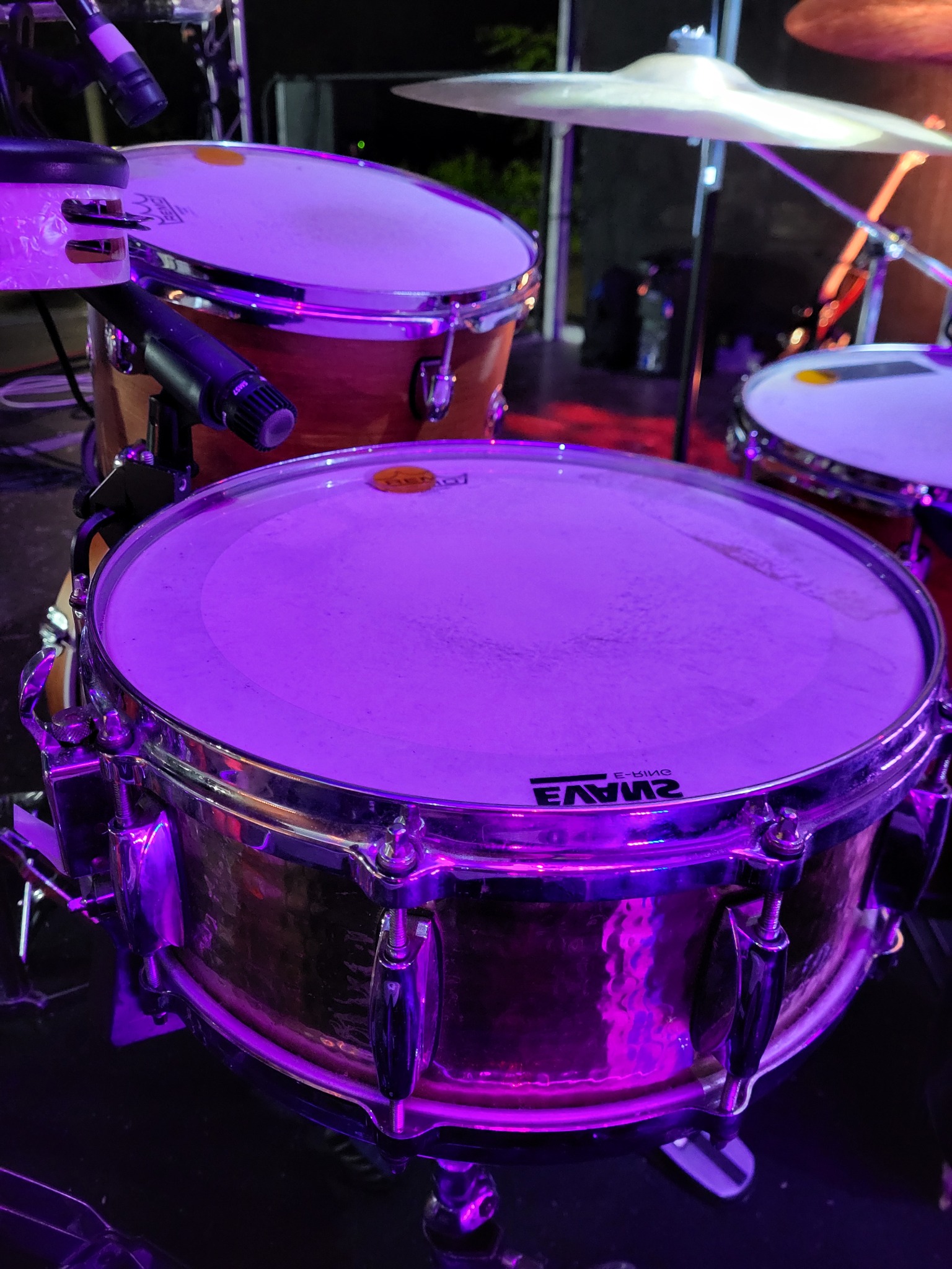 Perkusja z mikrofonami w fioletowym oświetleniu scenicznym. Widoczne bębny i talerze, gotowe do występu na żywo. Sprzęt muzyczny w blasku reflektorów.