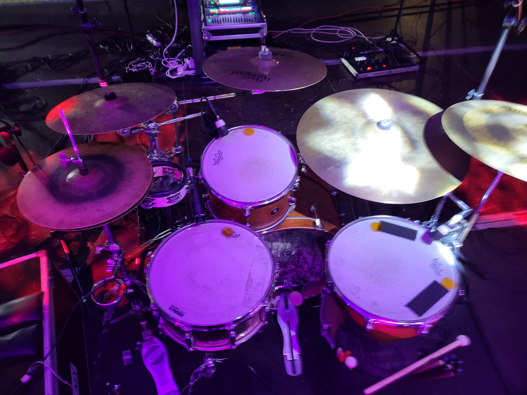 Zestaw perkusyjny z talerzami w fioletowym oświetleniu scenicznym, widok z góry. Na bębnie naklejone czarne i żółte tłumiki. Widoczne kable i sprzęt audio w tle.