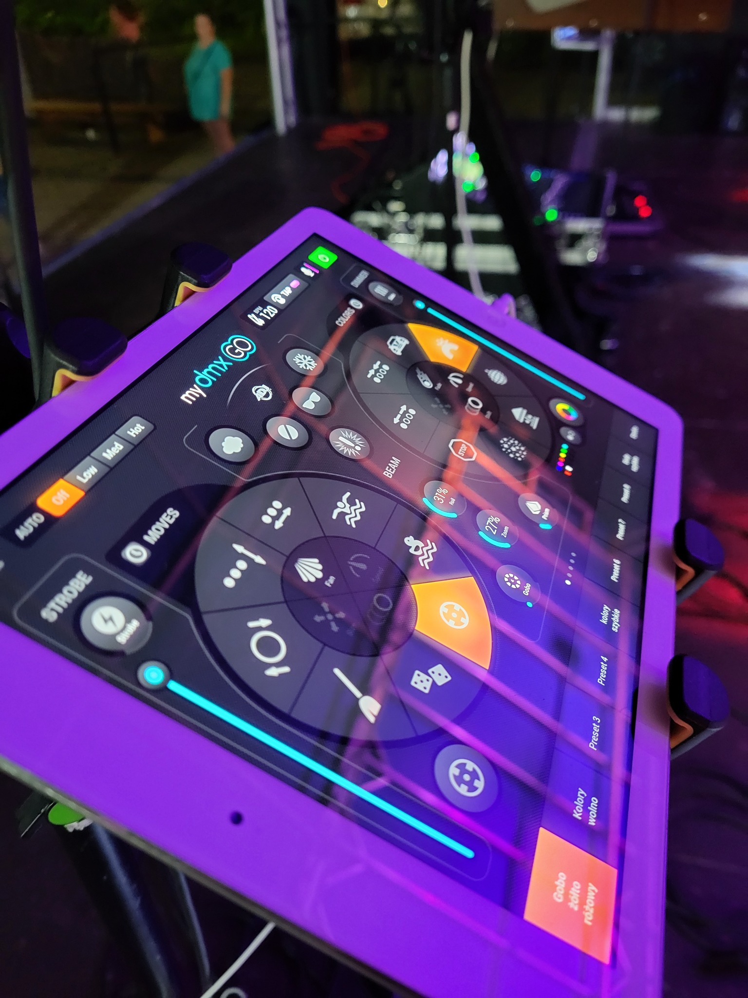 Tablet z aplikacją myDMX GO, sterujący oświetleniem scenicznym. Widoczne opcje: strobo, kolory, ruchy. W tle sprzęt oświetleniowy, ustawiony pod kątem.