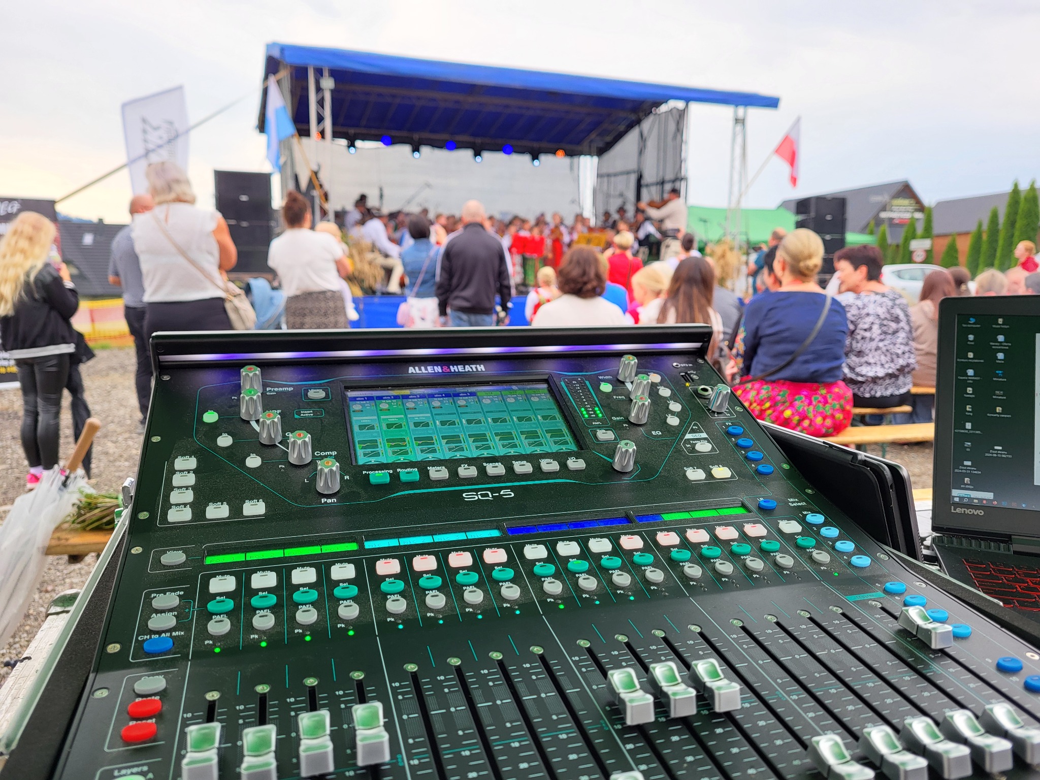 Konsoleta mikserska Allen & Heath SQ-5 na tle sceny plenerowej z występującymi osobami i publicznością. Widoczny laptop i elementy wyposażenia.
