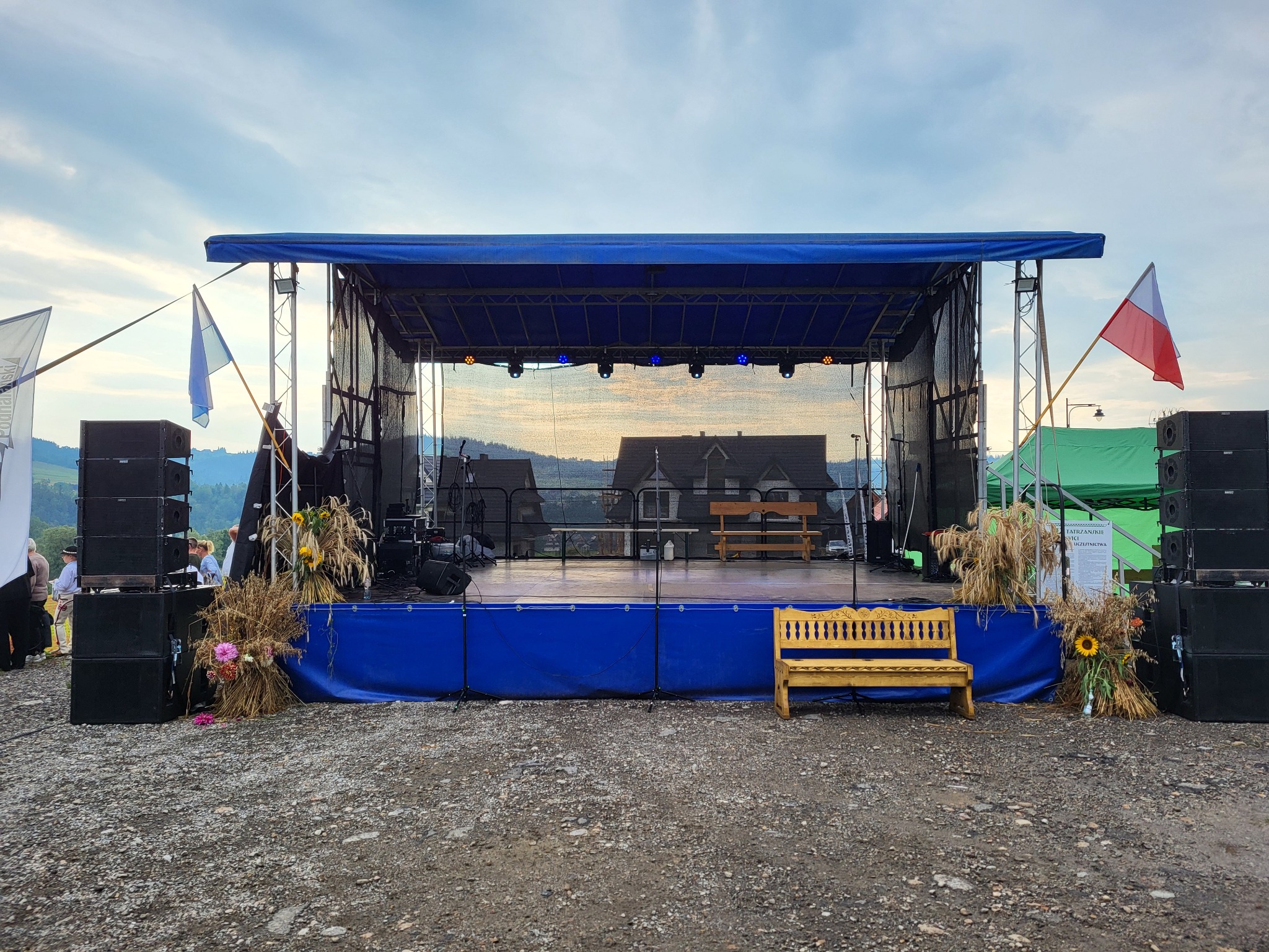 Scena plenerowa z nagłośnieniem i dekoracjami z kwiatów i zboża, w tle góry i domy. Flagi polska i niebieska po bokach sceny.