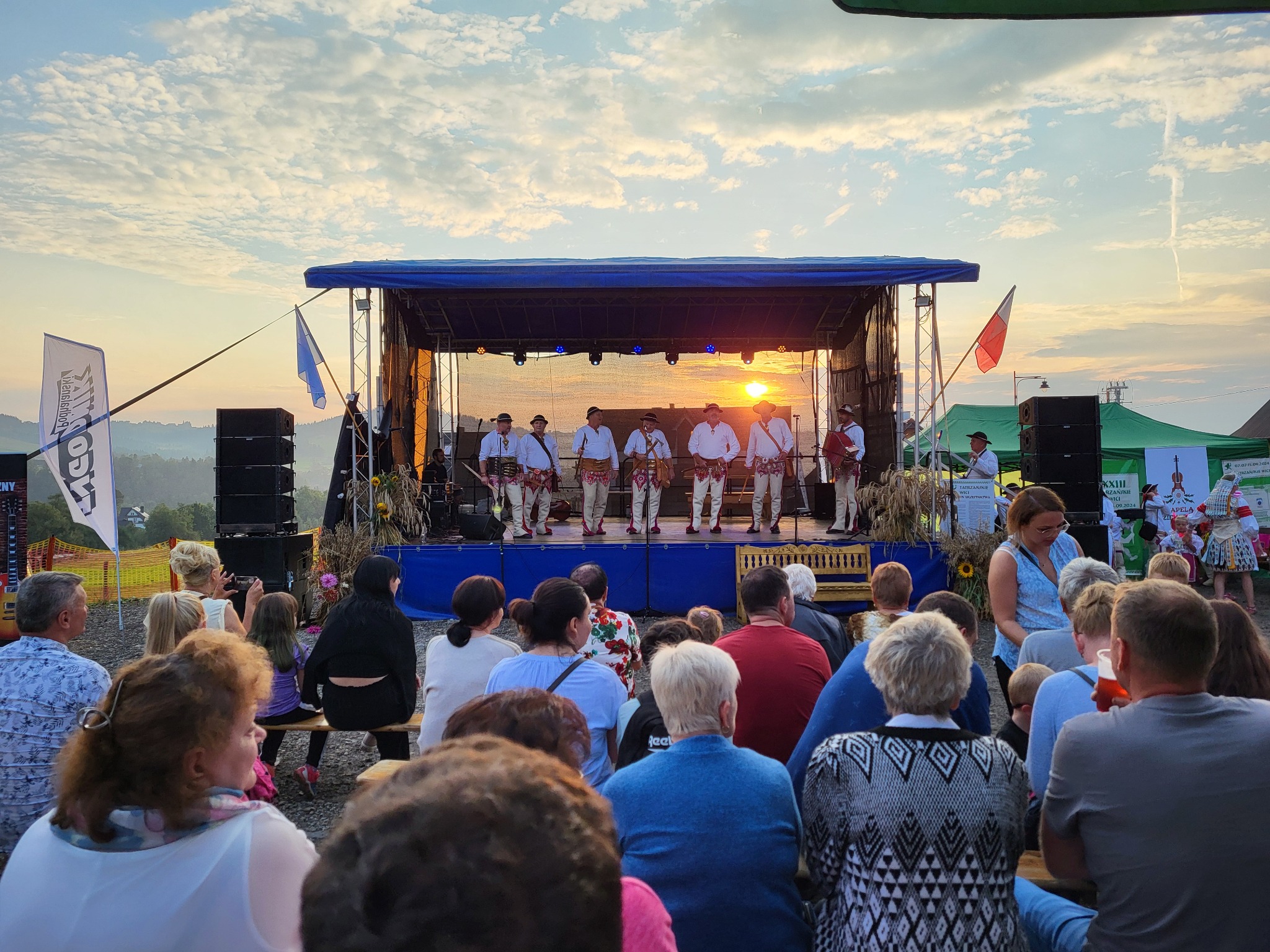 Zespół w strojach ludowych na scenie, grający koncert plenerowy dla publiczności siedzącej na ławkach. Zachód słońca w tle, flagi i nagłośnienie po bokach sceny.