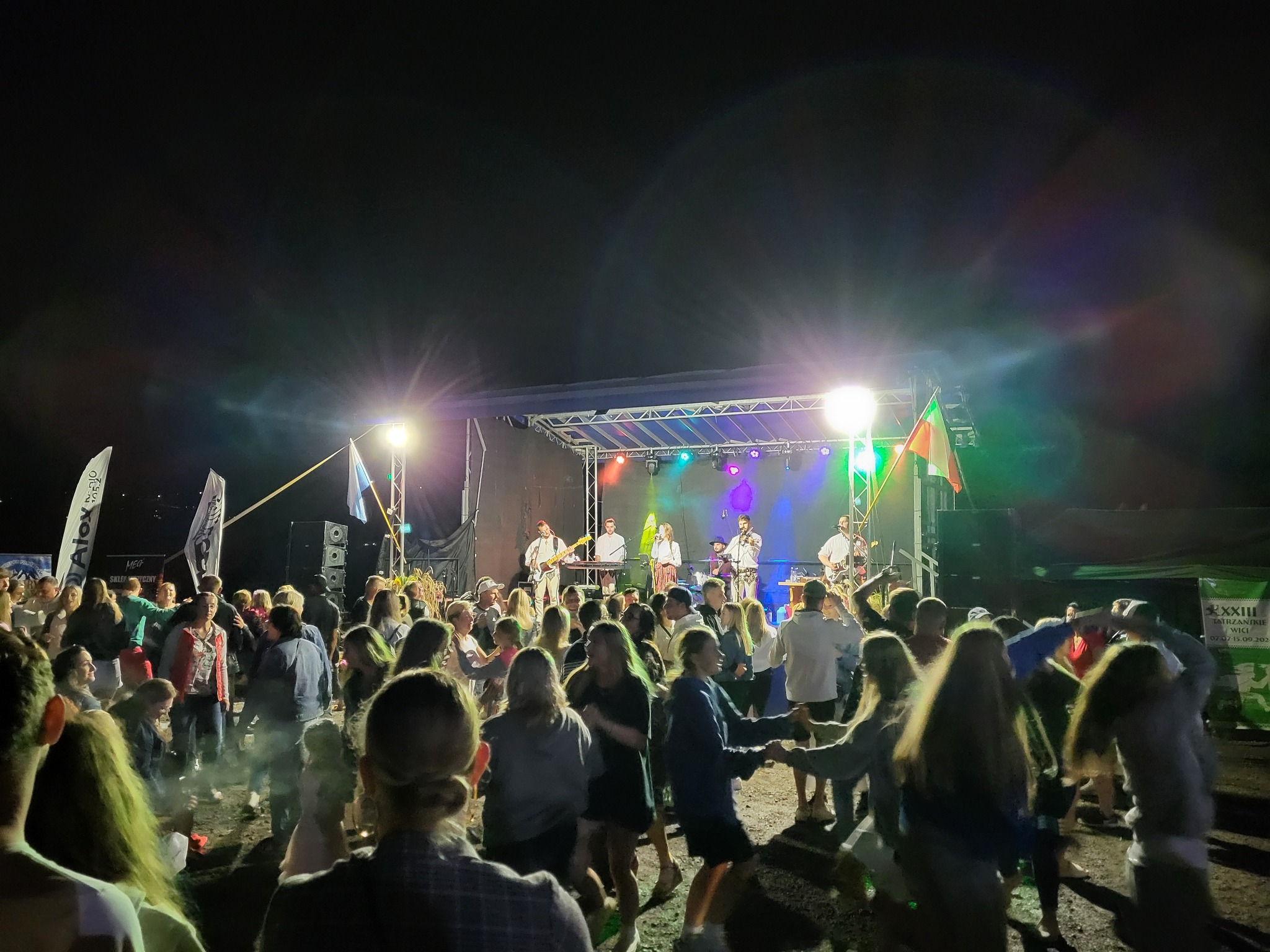 Zespół muzyczny na scenie gra koncert dla tańczącego tłumu w nocy. Widoczne oświetlenie sceniczne i flagi. Ujęcie z perspektywy widza.